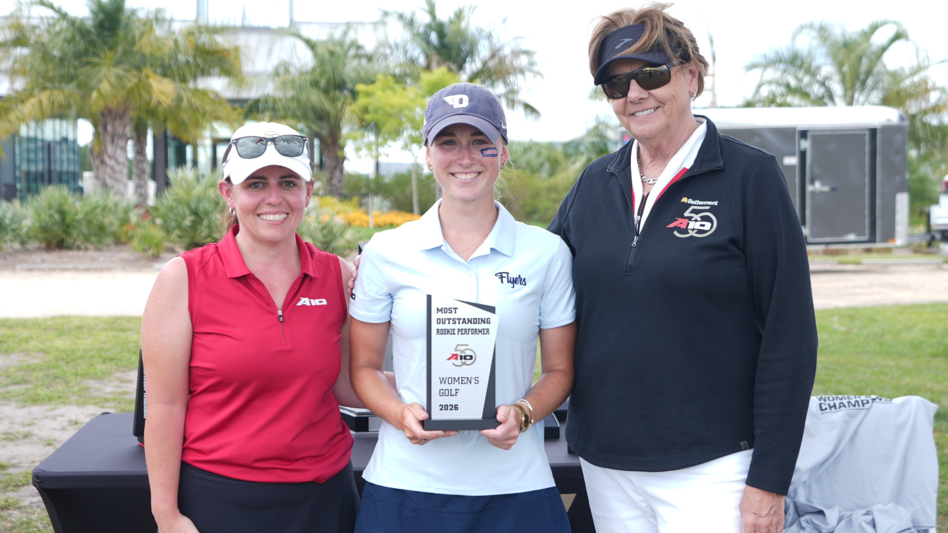 Women's golfer Etta Huffman, wearing a blue hat, white shirt and blue skirt, stands between two Atlantic 10 administrators holding the Atlantic 10 Most Outstanding Rookie Performer trophy following the conference tournament