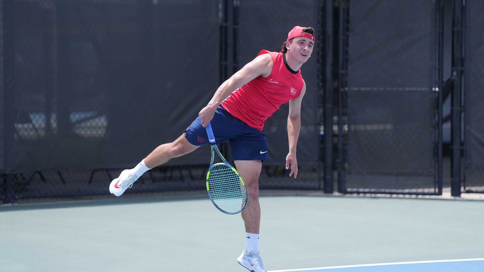 Dylan Jaen is on his left leg with his right leg out behind him after hitting the ball. He is wearing a red hat backwards with a red tank top and navy shorts.