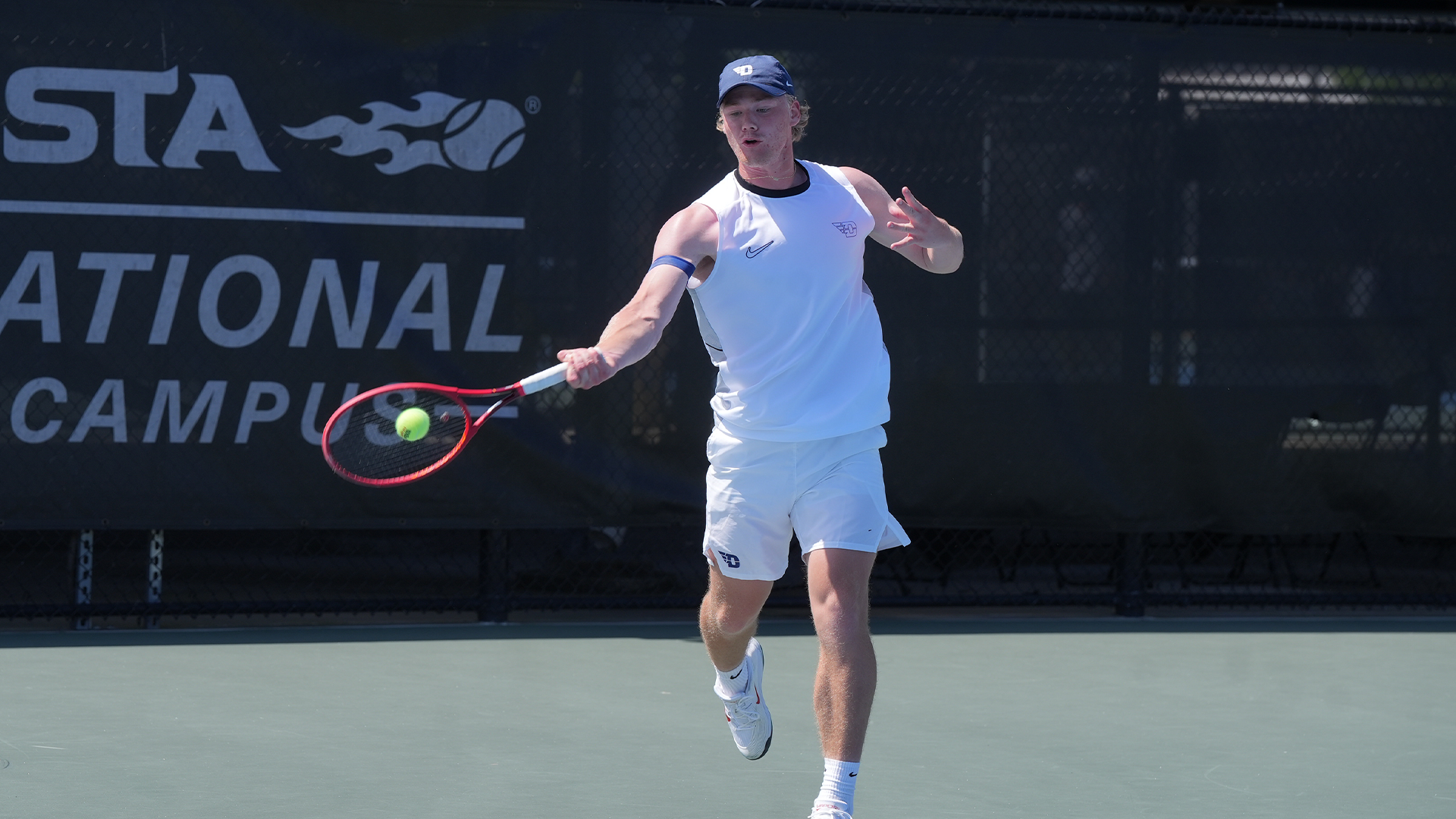 Sturle Skigelstrand, wearing a white tank top and shorts and a navy hat, hits the ball with a red racket in his right hand.