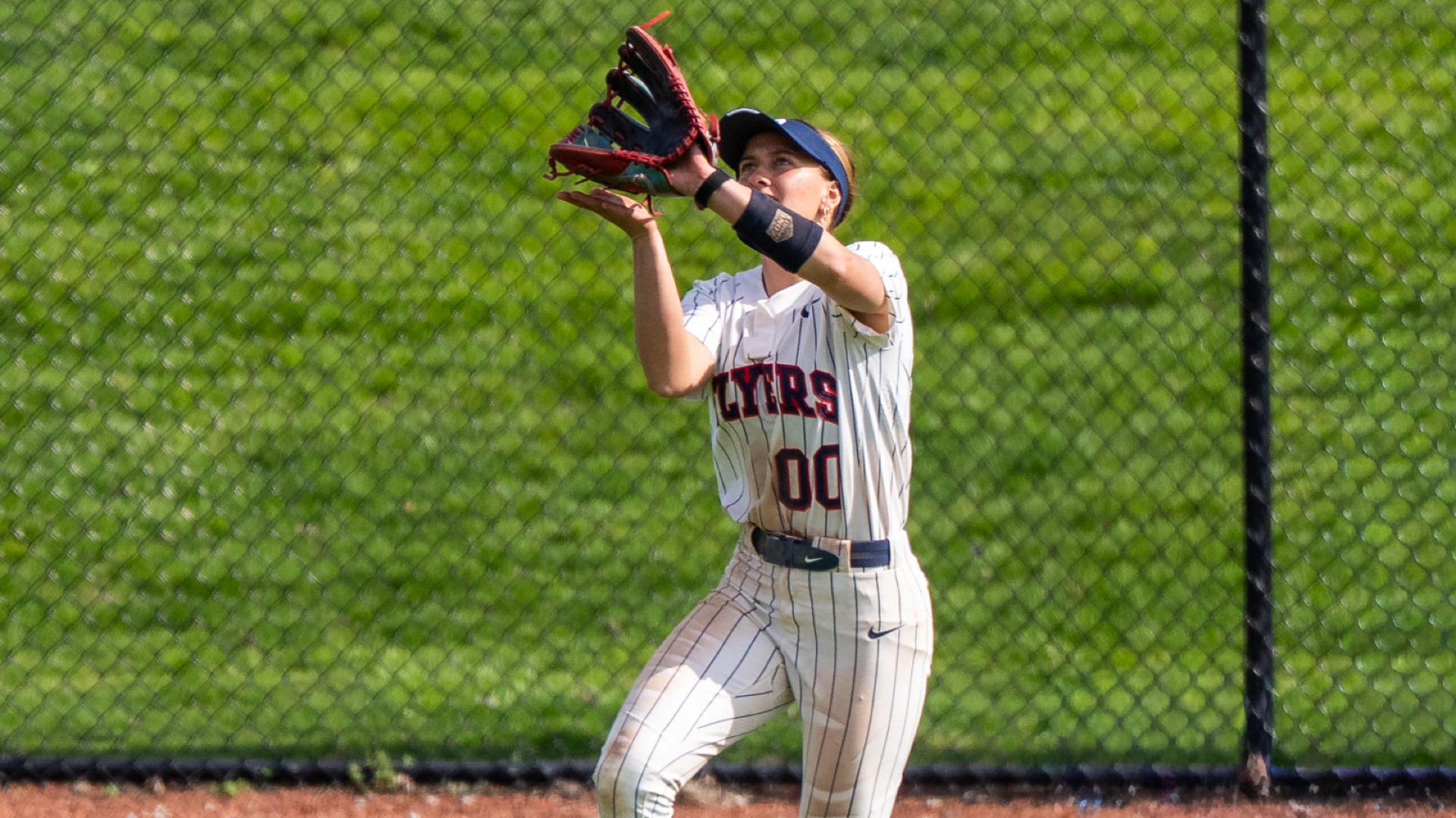 Softball's number 00 Camrynn Linneman, wearing a white pinstripe uniform and navy blue visor hat, prepares to catch a fly ball with her glove and right hand above her face ready to squeeze