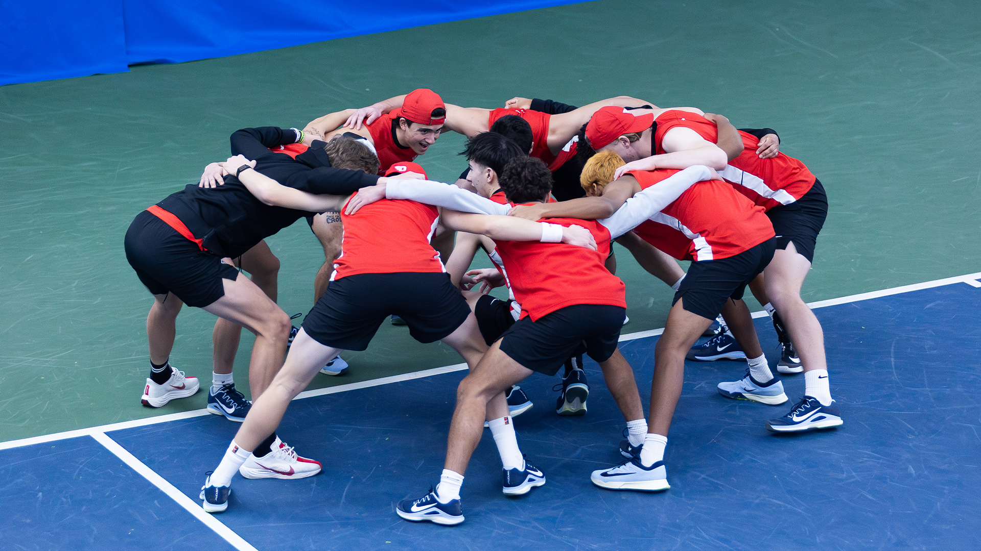 The men's tennis team, wearing red tank tops, huddle together before the match.