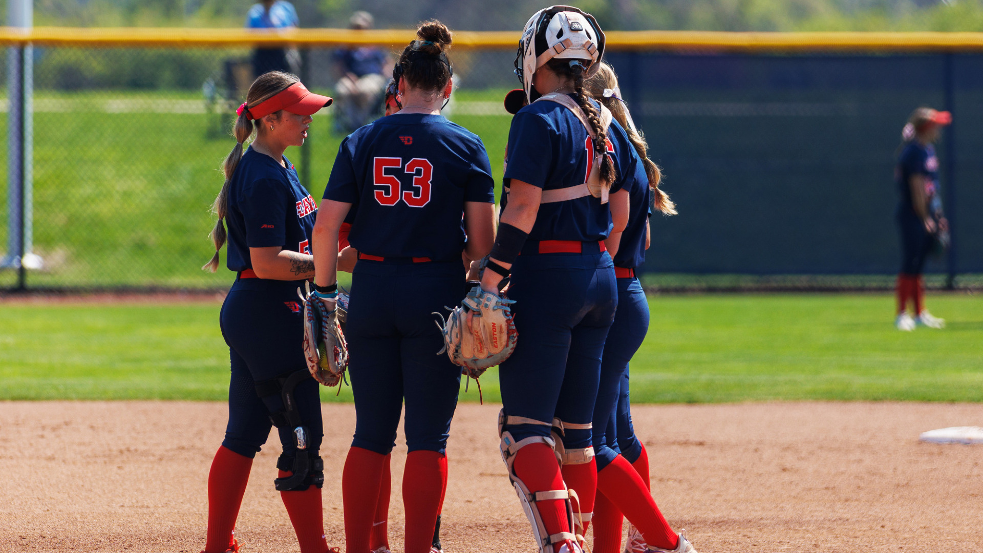Members of the softball infield, wearing blue uniforms with red numbers, gather in the pitcher's circle to talk it over during a game at UD Softball Stadium