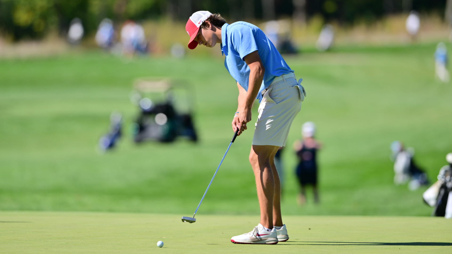 Men's golfer Victor Caliguri, wearing a light blue polo-style shirt, khaki shorts and a red and white trucker-style hat, puts on a green at Moraine Country Club