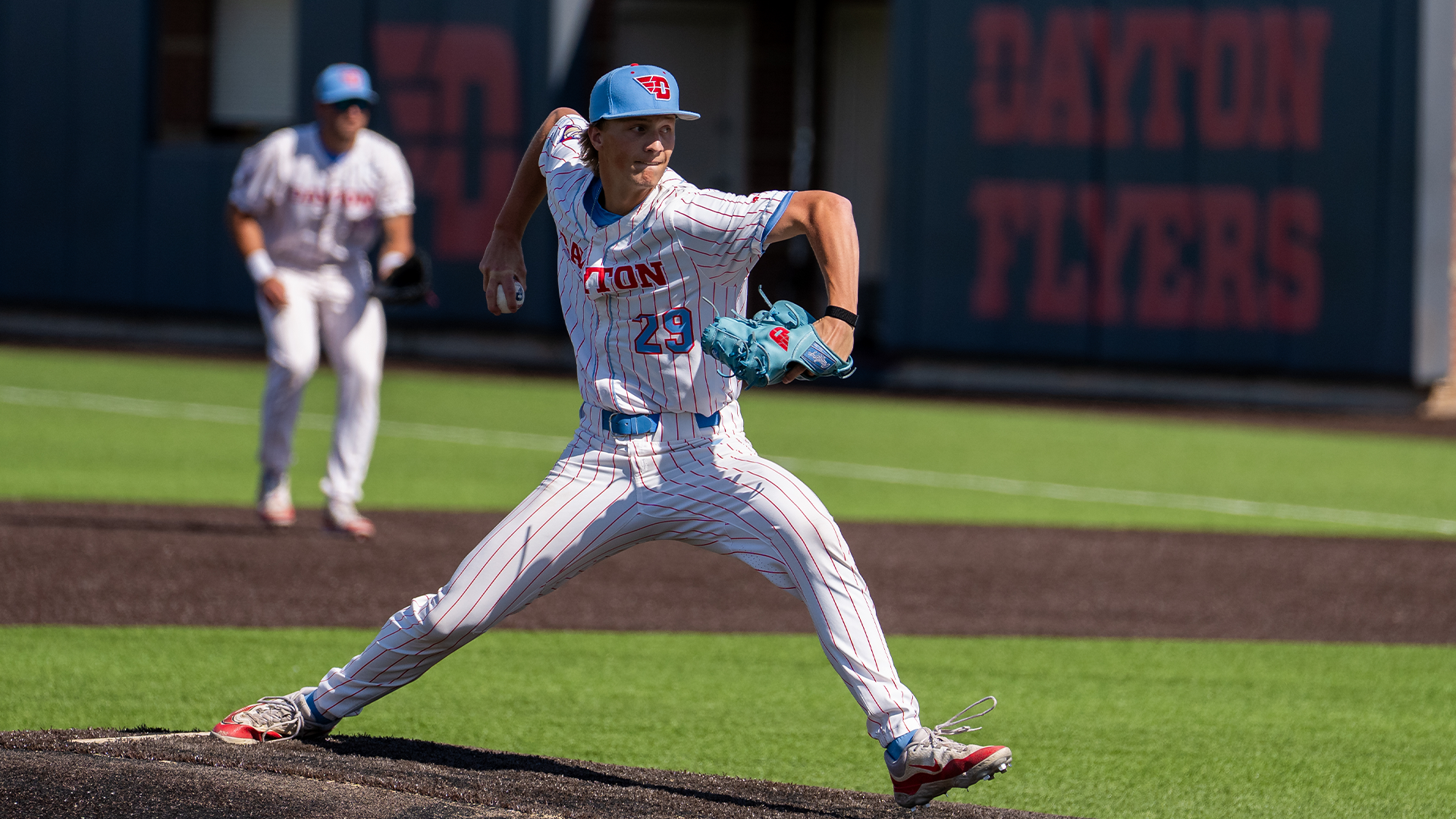 Austin McNabb has his right arm wound back as he throws the pitch. His left leg is stretching forward. He is wearing a white pinstripe uniform and a light blue hat.