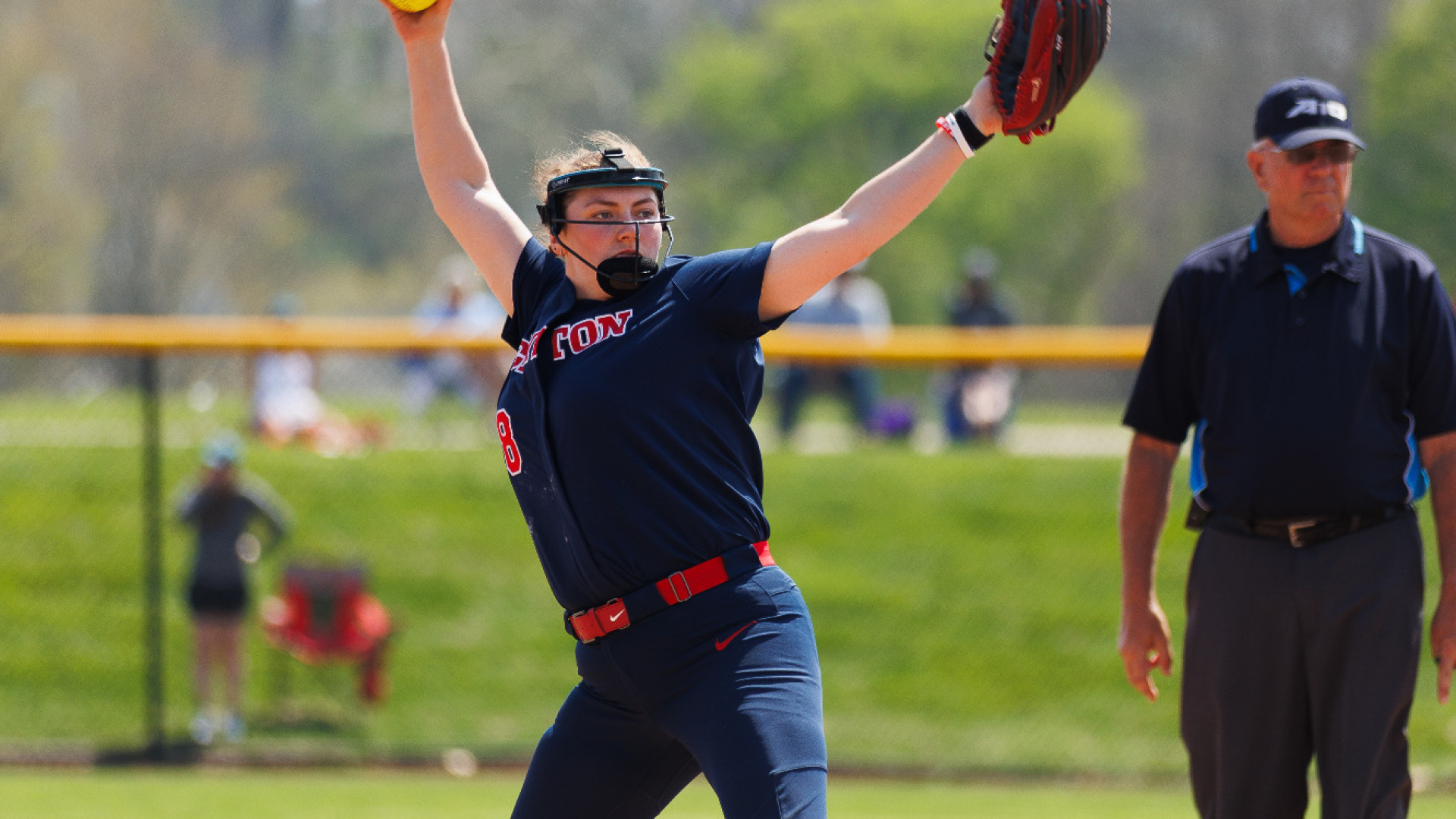Softball pitcher Kate Kastelic, wearing a blue uniform top and bottom, red socks and protective face gear prepares to throw a pitch in a game at UD Softball Stadium