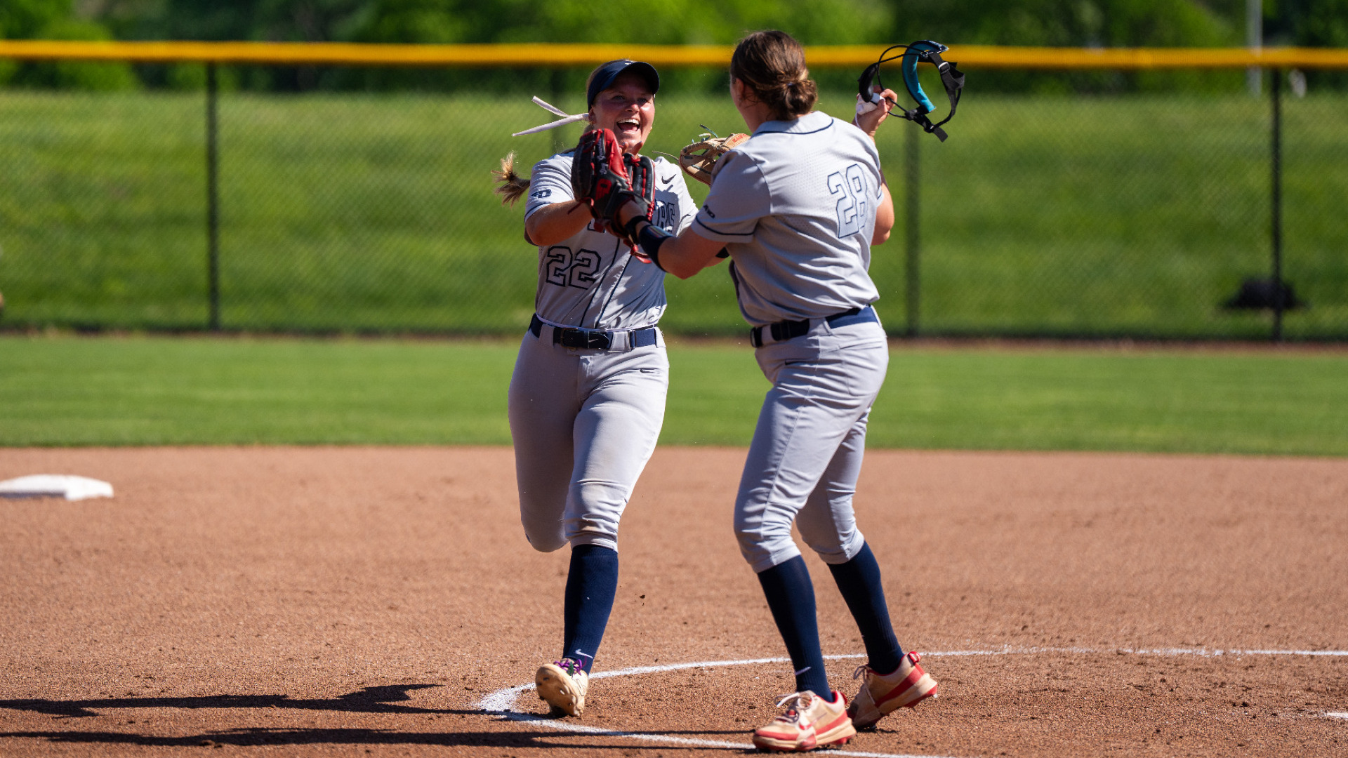 Softball players number 22 Molly Grace and number 28 Kate Kastelic, wearing grey uniforms with blue socks, celebrate near the pitching circle during a game at UD Softball Stadium