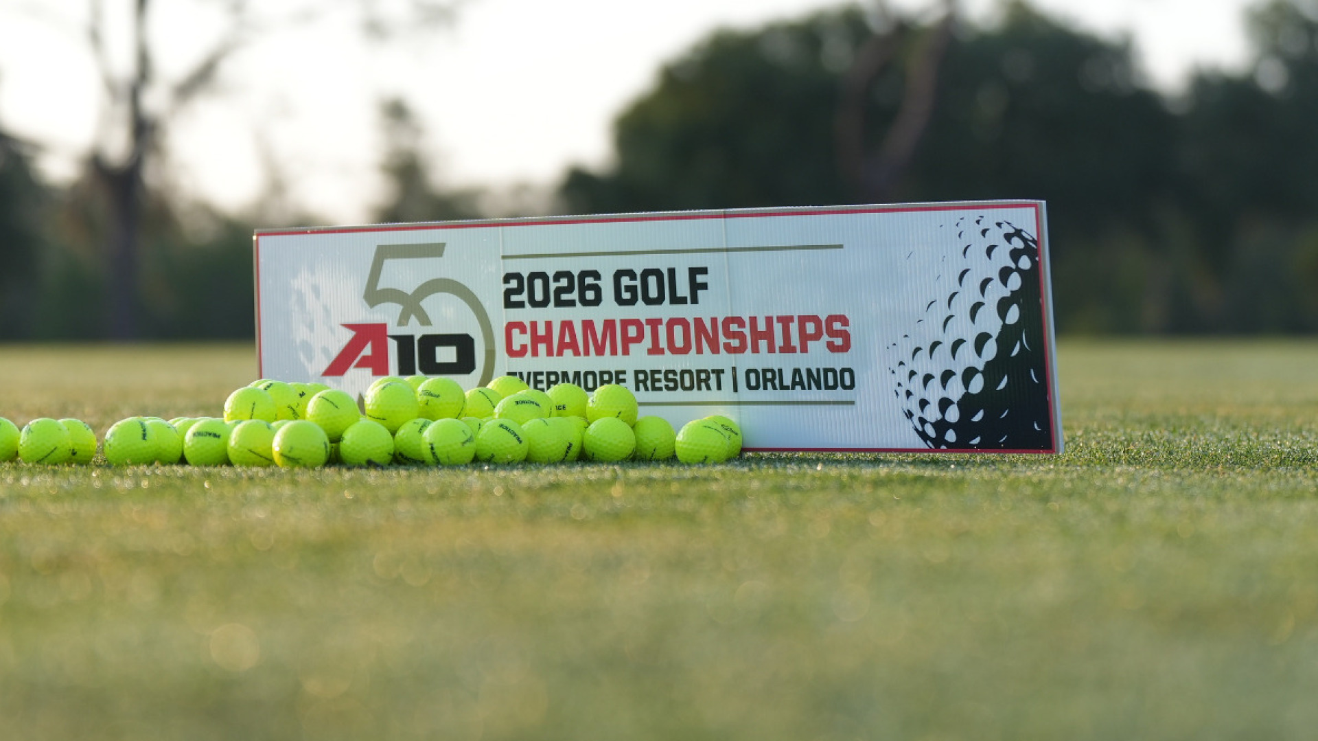 A sign with an Atlantic 10 50th anniversary logo that reads 2026 Golf Championships Evermore Resort Orlando with several yellow range balls to the right of it