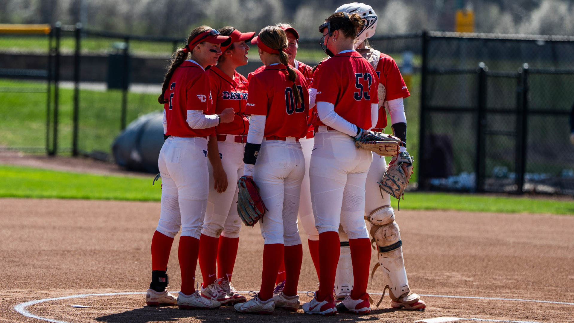 Members of the softball infield, wearing red uniform tops and white pants, gather in the circle for a huddle during a game at UD Softball Stadium