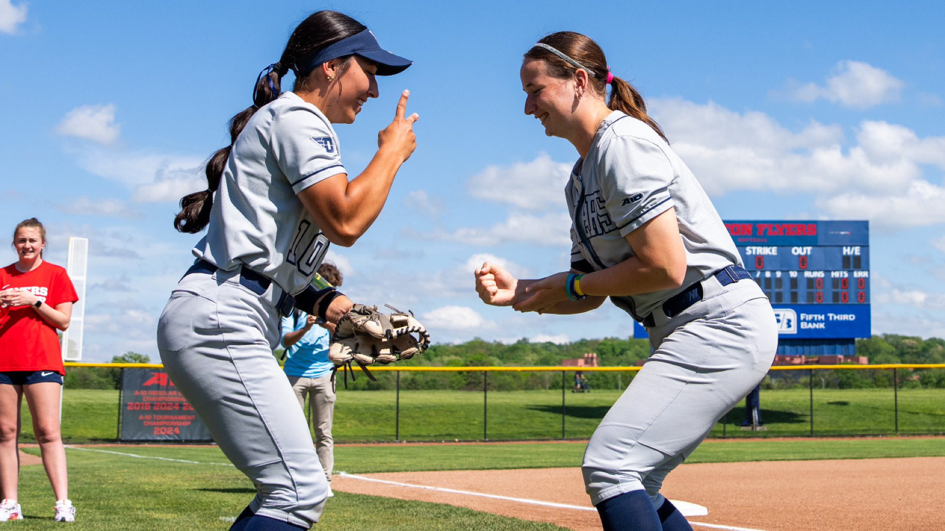 Softball players number 10 Izzy Calinski (left) and number 53 Izzy Kemp (right), wearing grey uniforms and blue socks, playing rock-paper-scissors during starting lineups at a game at UD Softball Stadium