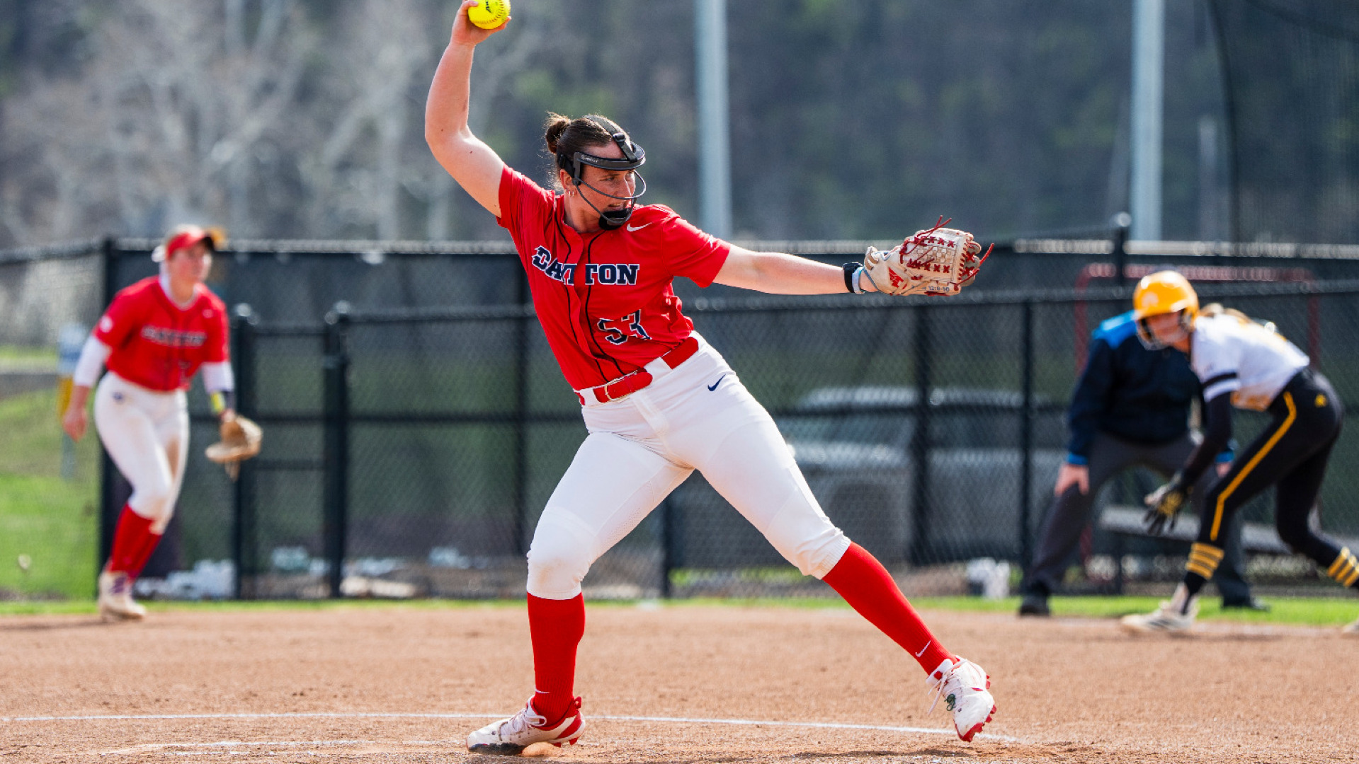 Softball pitcher Izzy Kemp, wearing a red Dayton uniform top with the number 53, white pants and red socks, with her hand above her head holding a ball in the top of her pitching motion in the circle