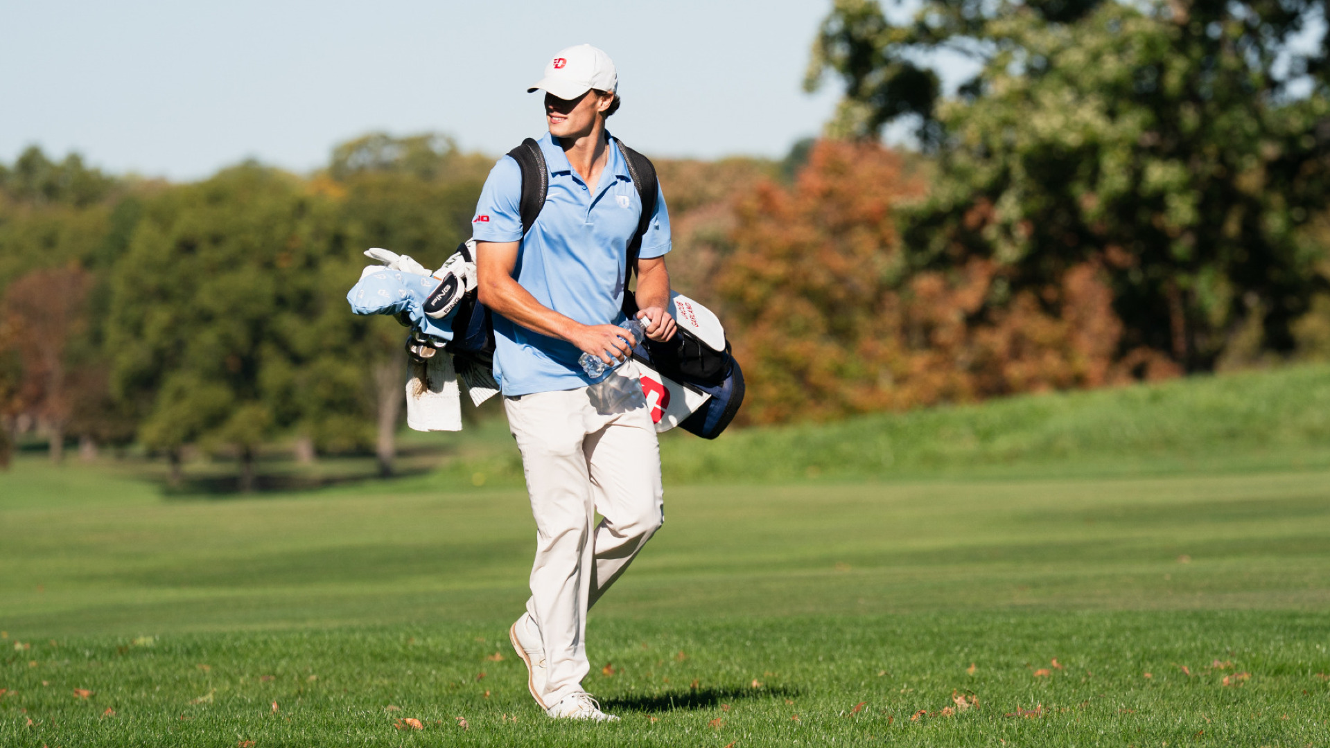 Men's golfer Jacob Garland wearing a chapel blue polo-style shirt, khaki pants and a white hat, walking down the fairway carrying his clubs, looking to his right and smiling