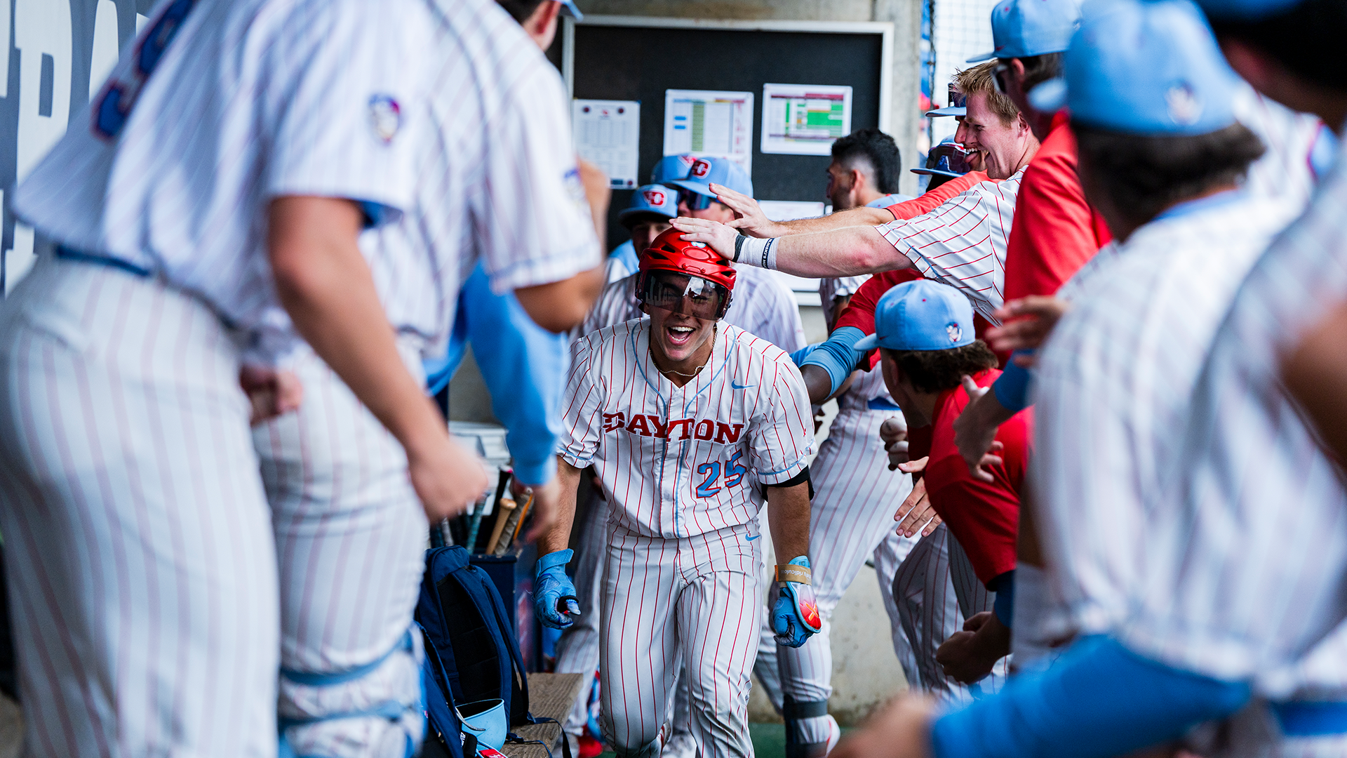 Grayson Carpenter, wearing a red hockey helmet, celebrates with the team in the dugout after a home run.