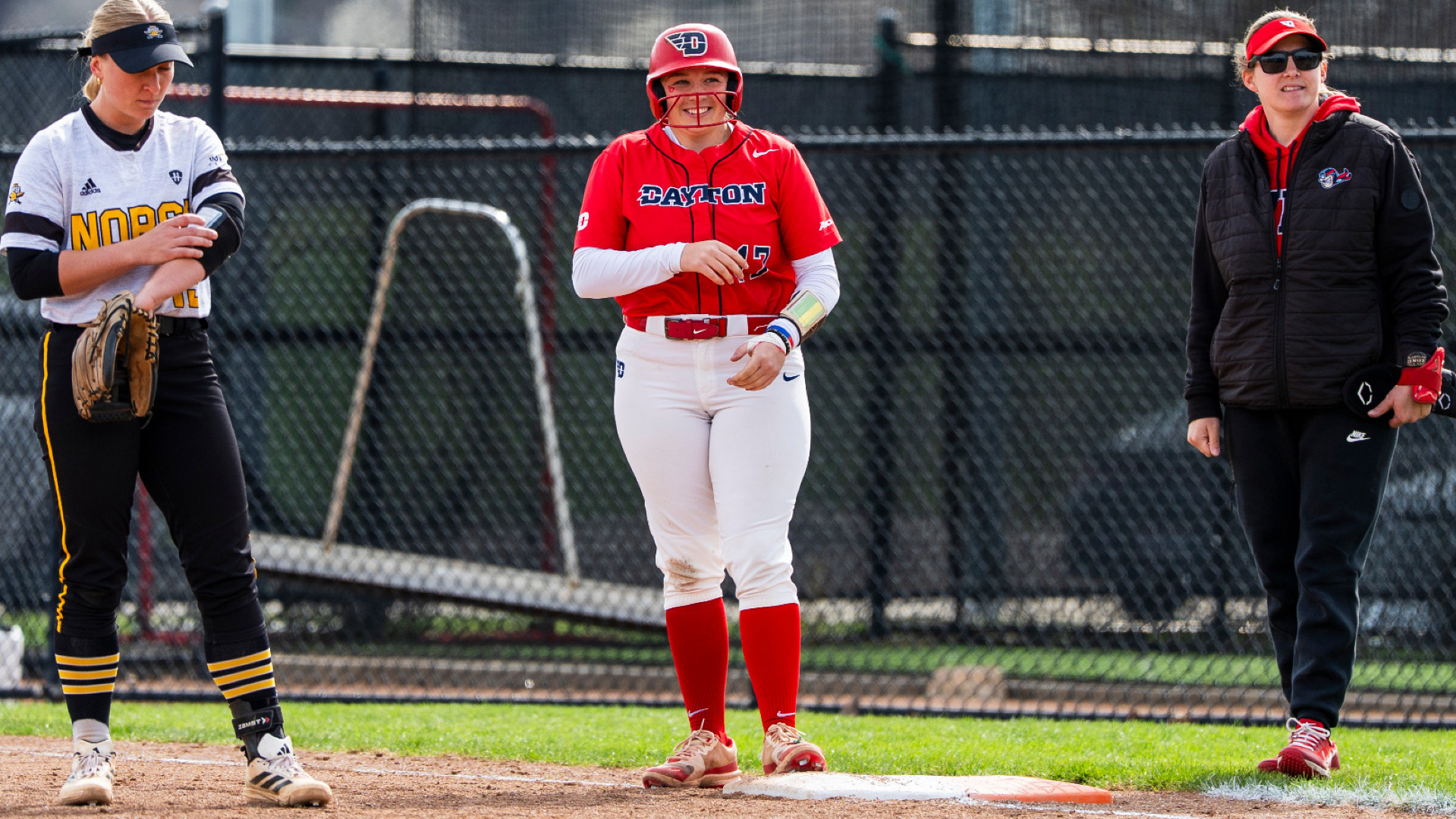 Softball player Deirdre Flaherty, wearing a red uniform top with the number 17 and white pants, stands on first base clapping