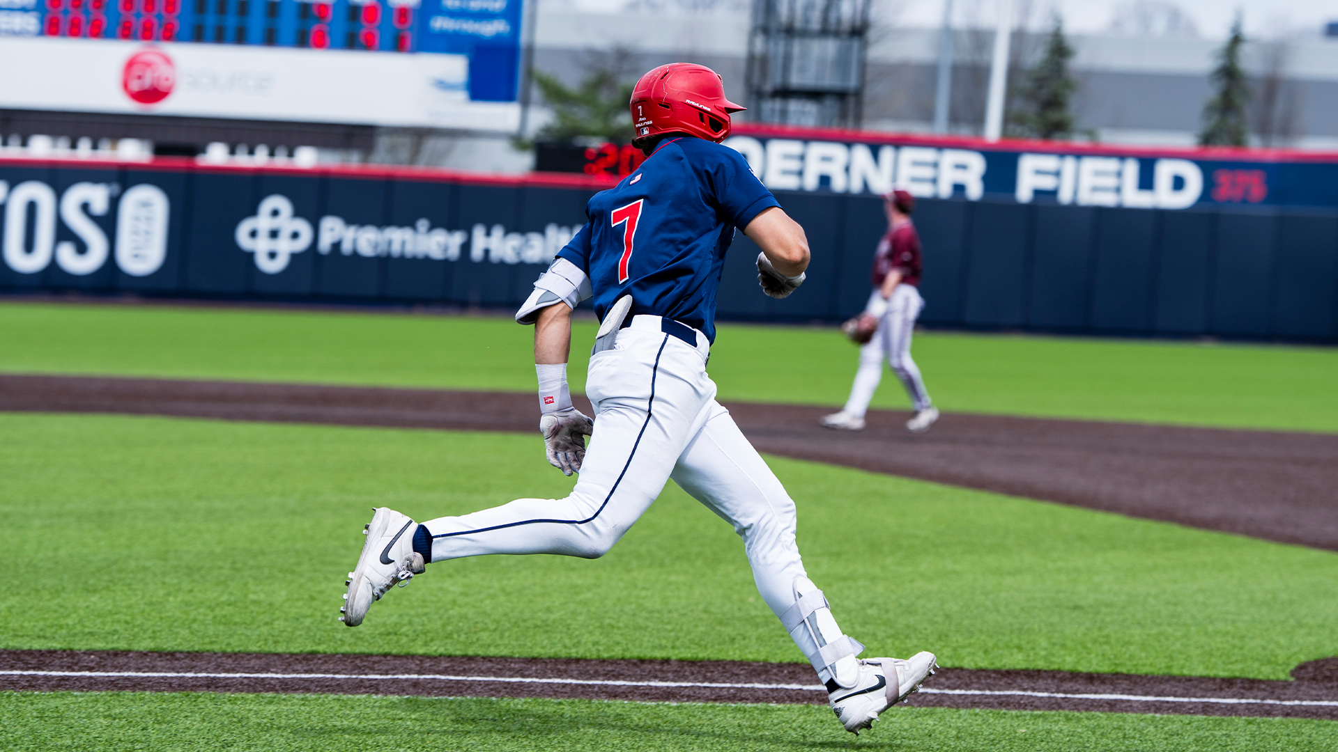 Jason Bello, wearing a navy jersey, white pants and a red helmet, runs to first base.