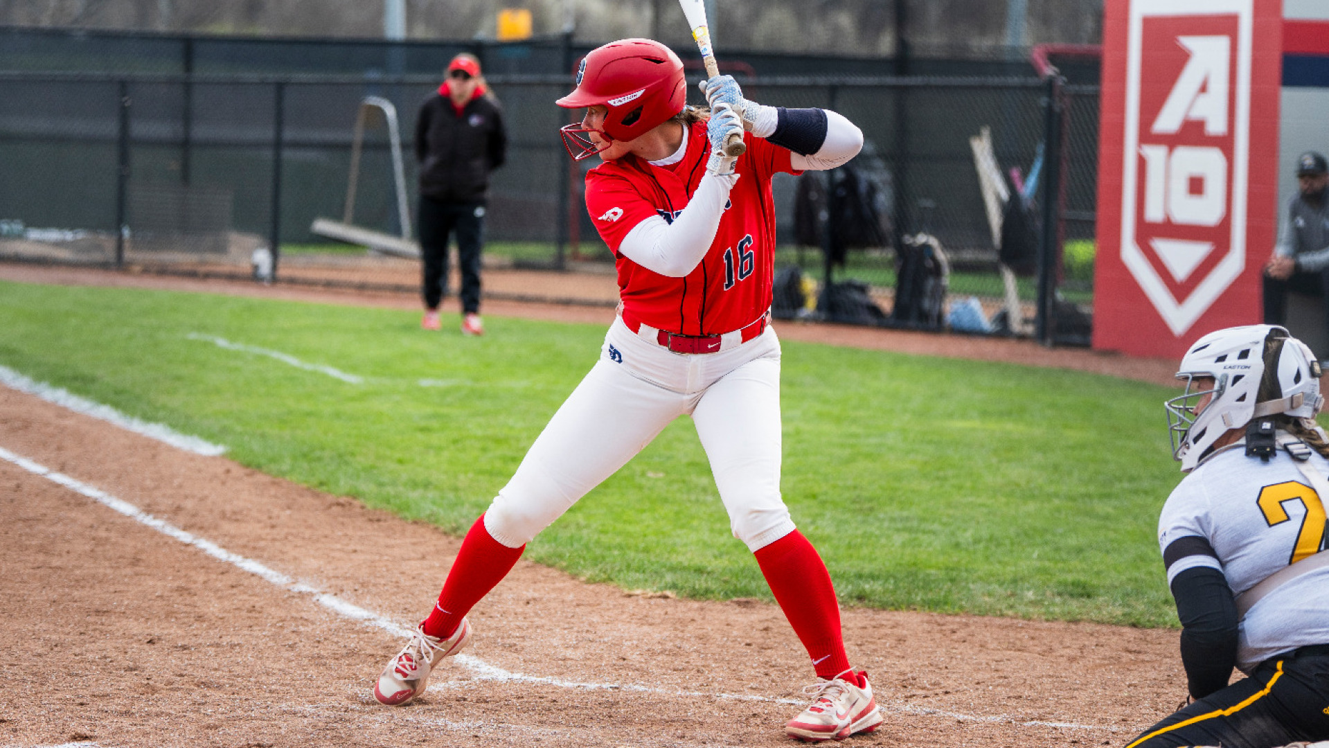Softball's Katelin Goodwin, wearing a red uniform top with the number 16, white pants and a red batting helmet, stands inside the box with a bat on her shoulder awaiting a pitch