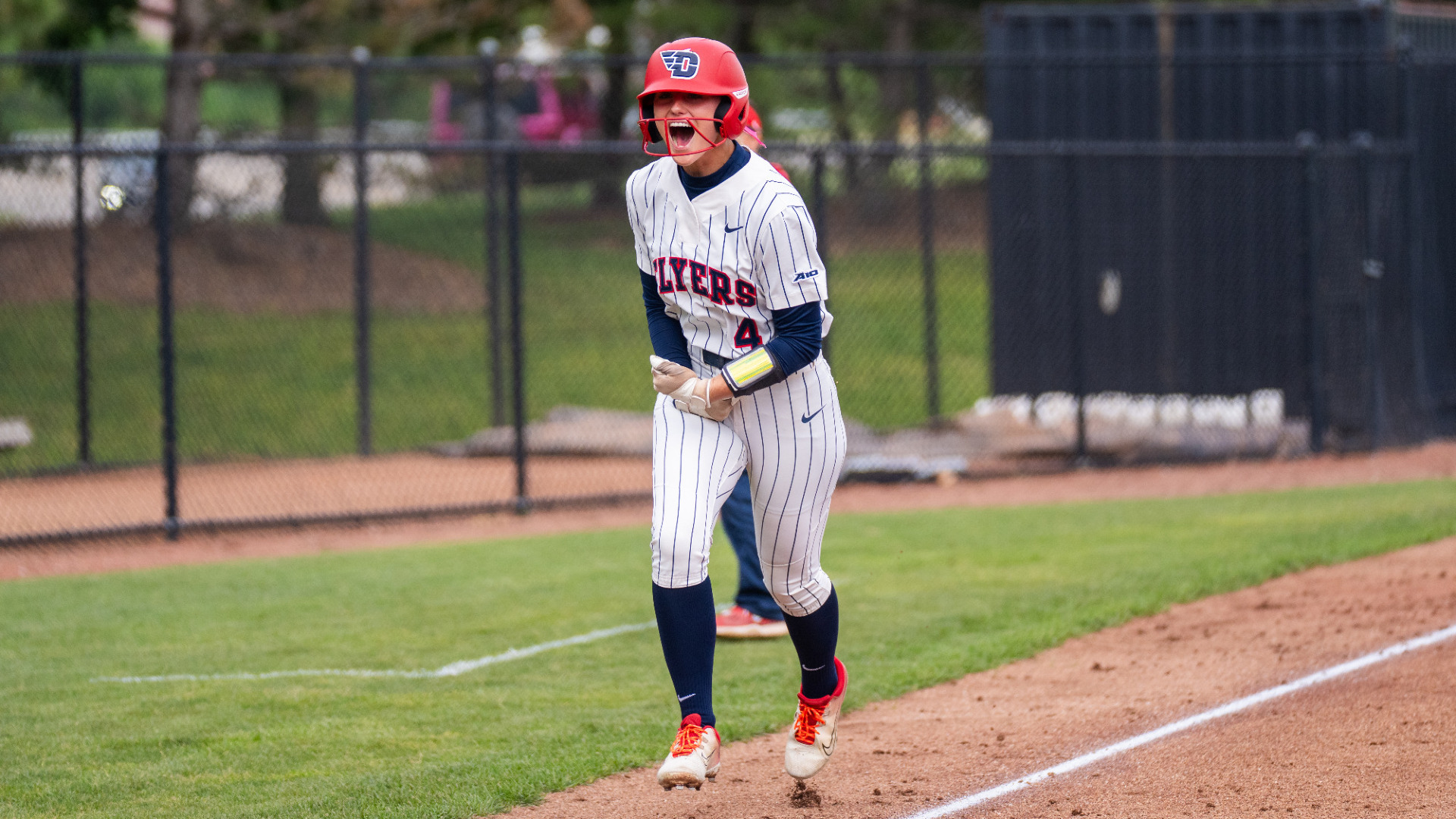 Softball's number four Mickey Shively, wearing a white pinstripe uniform and red batting helmet, running from third base to home with cheering after hitting a home run during a game at UD Softball Stadium