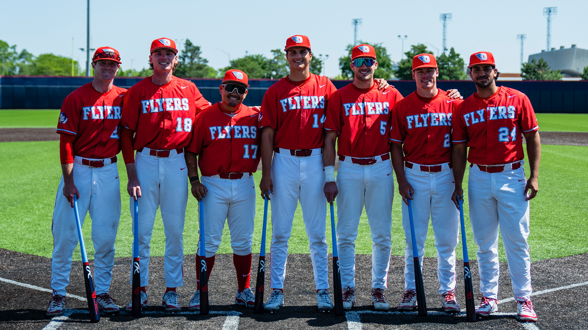 Graduating players Danny MacDougall, Jackson Judy, Dyrenson Wouters, Peyton Malecha, Aiden Heberlie, Tommy Ahlers Jr. and Michael Fortes pose for a picture. They are holding custom bats and wearing red jerseys with white pants.