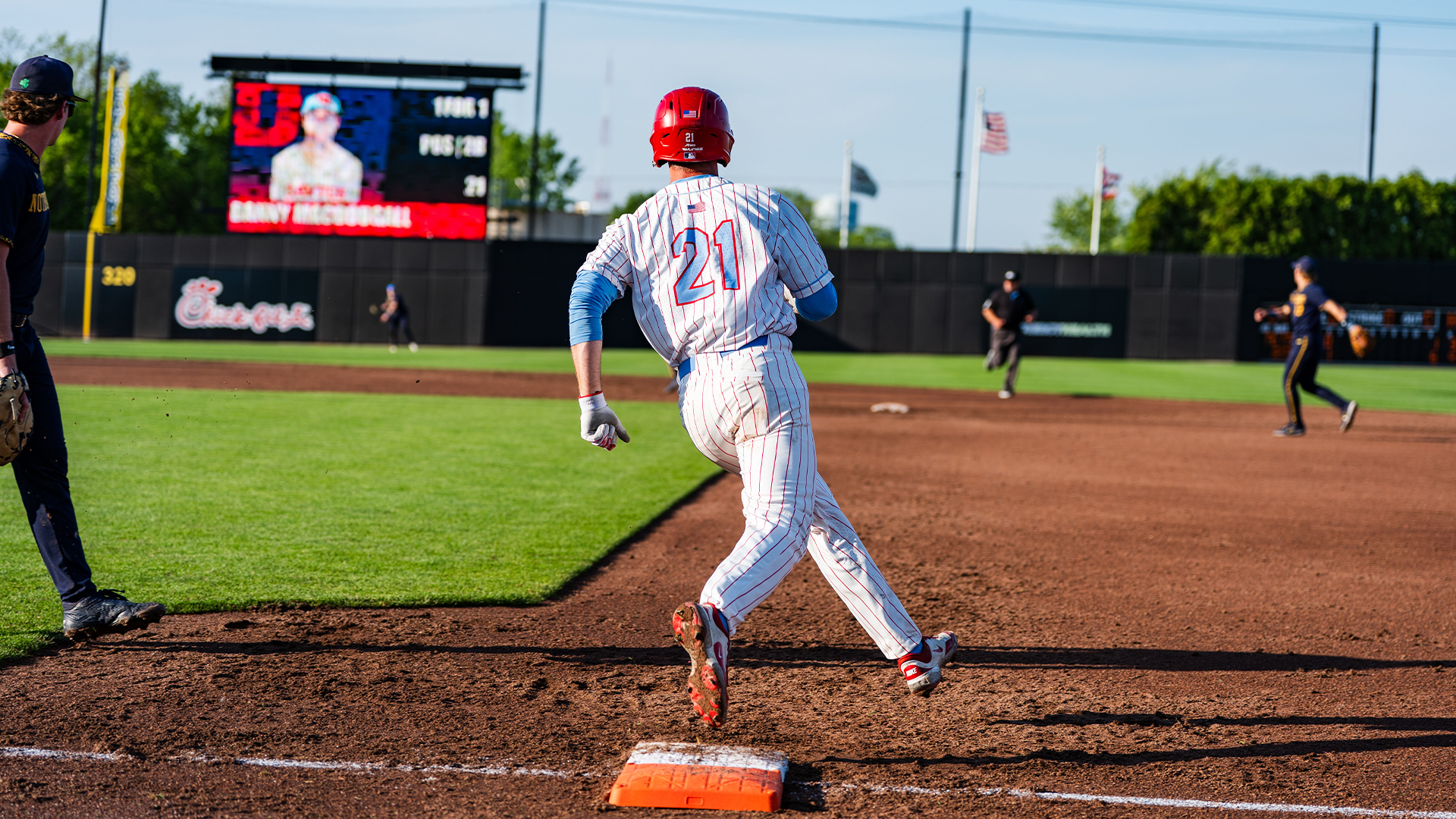 Danny MacDougall rounds first base at Prasco Park. His back is to the camera. He is wearing a white pinstripe uniform with a red helmet.