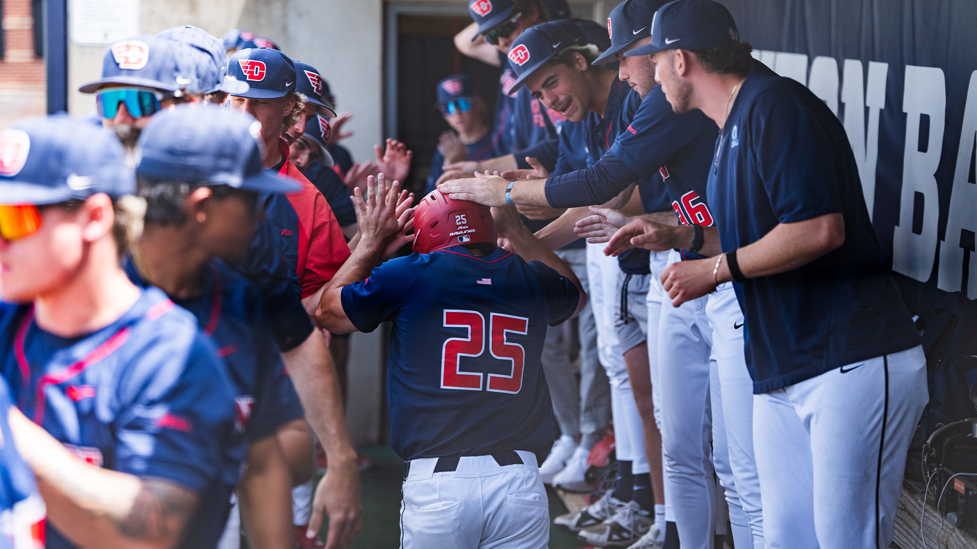 Grayson Carpenter runs through the dugout, high-fiving and getting hit on the helmet by teammates. He is running away from the camera, so his back is to the viewer. The team is wearing navy jerseys and white pants.