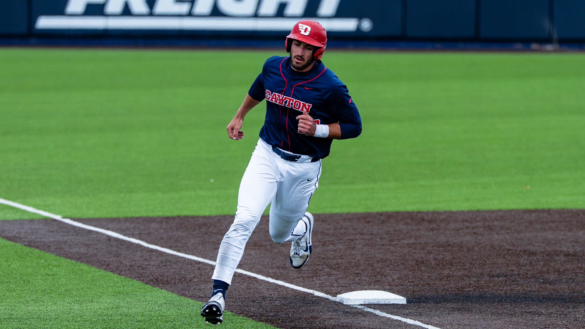 Bobby Stang, wearing a red helmet, navy jersey and white pants, rounds third base towards home.