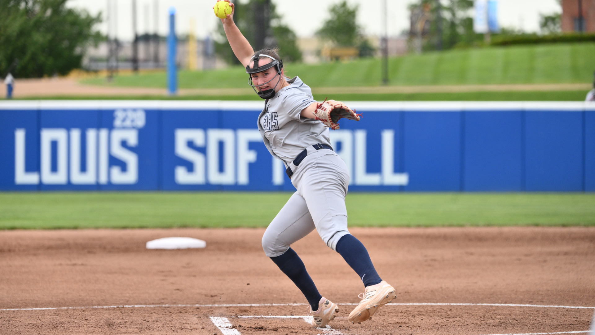 Softball pitcher Izzy Kemp, wearing a gray uniform, navy socks and protective face gear, is at the top of her throwing motion with her gloved left hand in front of her