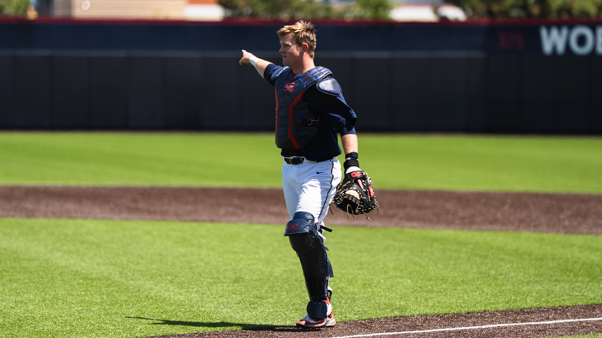 Ryan MacDougall, wearing his catcher's gear, a navy jersey and white pants, stands on the first base line and points to the infield.
