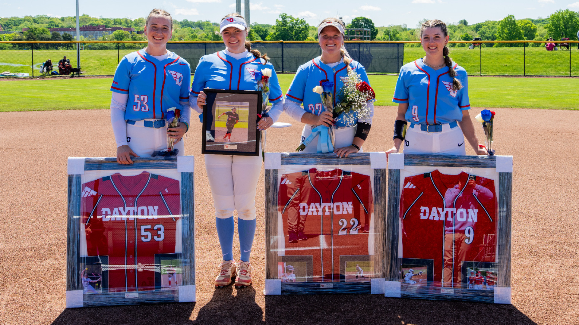 From left to right, Izzy Kemp, Kate Kastelic, Molly Grace and Madelyn Fricano, wearing a light blue uniform top with white pants, are holding frames and flowers following senior day recognition at UD Softball Stadium