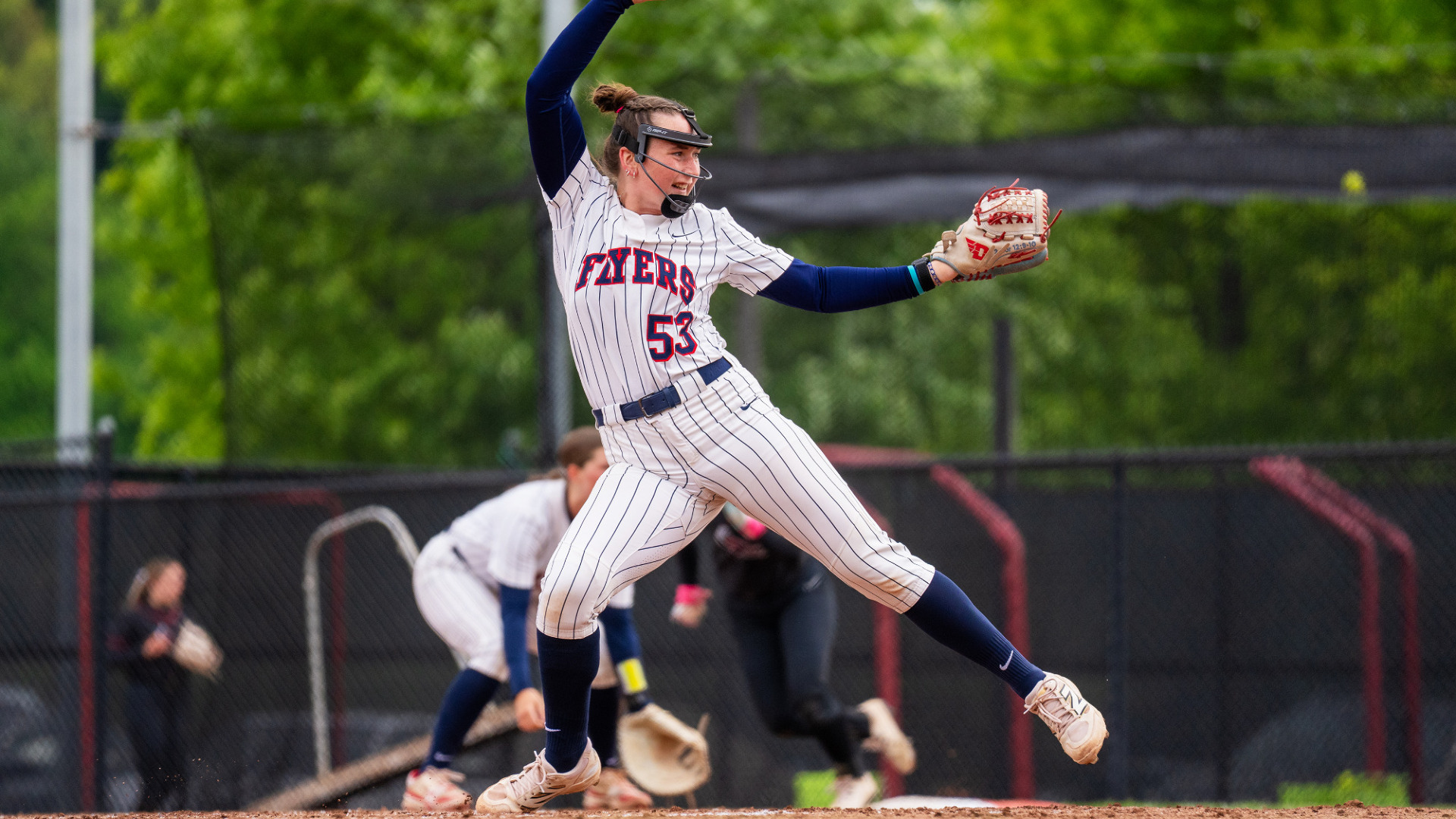 Pitcher number 53 Izzy Kemp in the circle at UD Softball Stadium, wearing a pinstripe uniform, blue socks and protective face gear, has her had above her head at the top of her throwing motion