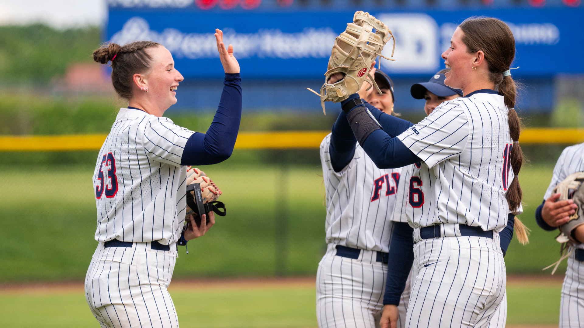 Softball number 53 (left) number 16 (right), wearing white pinstripe uniforms, high five near the pitcher's circle gathered with other members of the infield