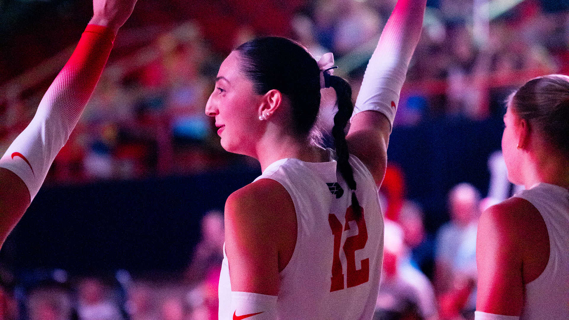 Liana Sarkissian stands with her teammates during starting lineups, holding her right arm in the air and pointing. She is wearing a white uniform with red details.