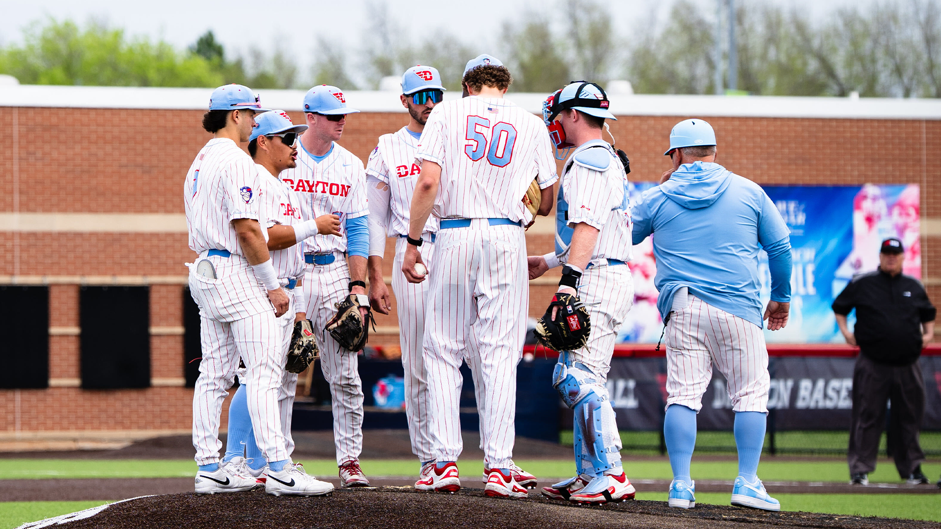 The infield players huddle on the mound with Coach King. The players are in a white pinstripe uniform. Coach King is wearing white pinstripe pants and a chapel blue sweatshirt.