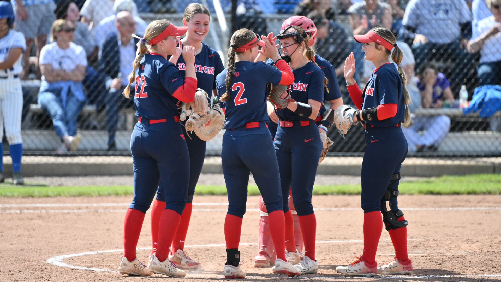 Softball infielders, earing navy blue uniforms with red numbers and red socks, gather in the circle during a game at Saint Louis's Billiken Sports Center