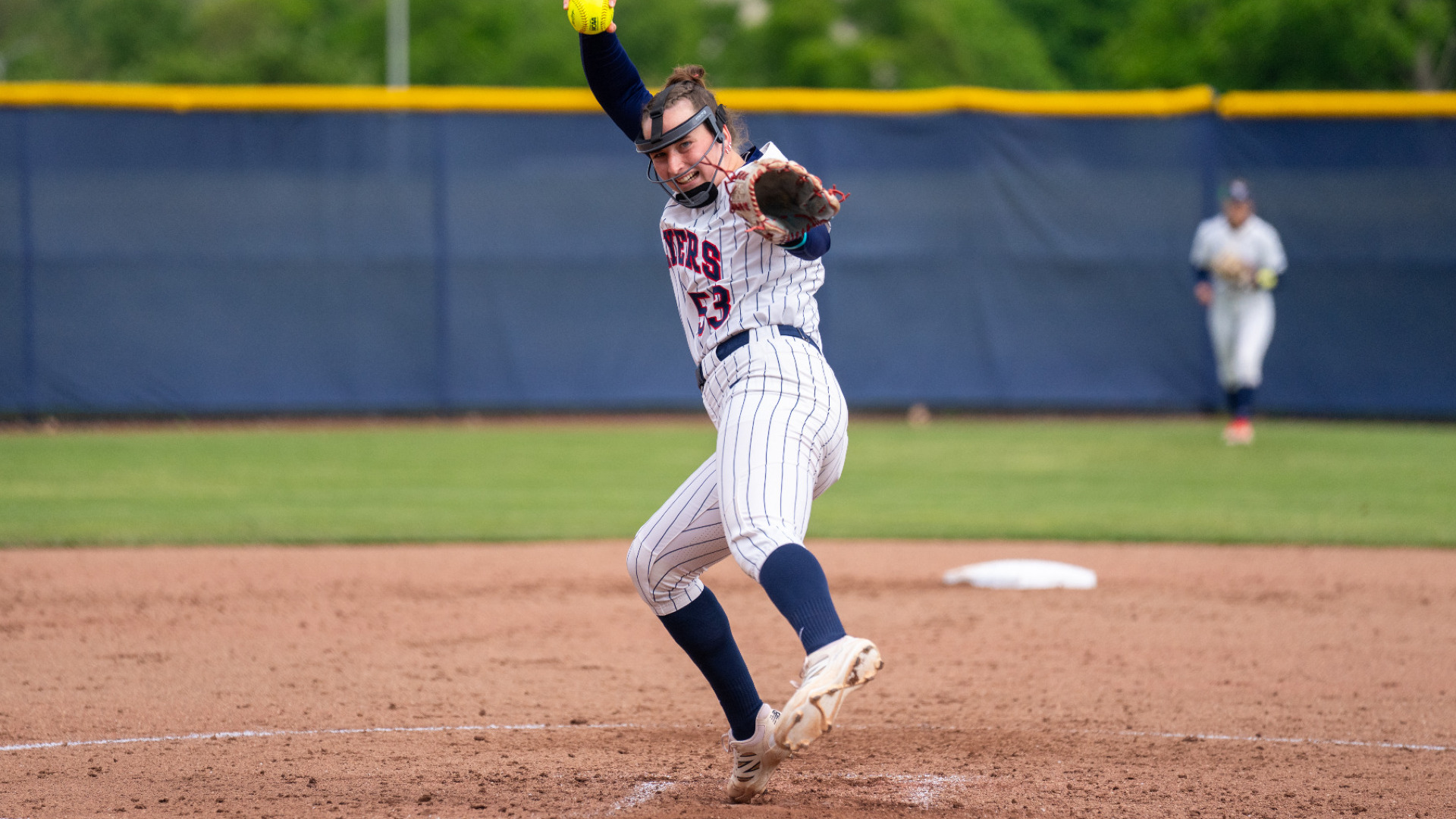Softball's number 53 Izzy Kenmp, wearing a white pinstripe uniform with navy socks and a protective face mask, has a ball above her head at the top of her throwing motion and her gloved left hand out in front of her mid pitch during a game at UD Softball Stadium