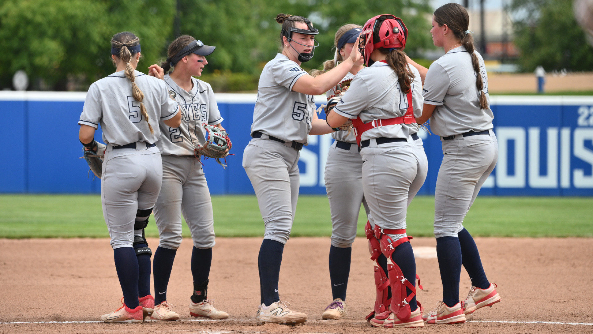 Members of the softball infield gather in the circle before the start of an inning, wearing gray uniforms with navy socks, fiving each other before heading into the field