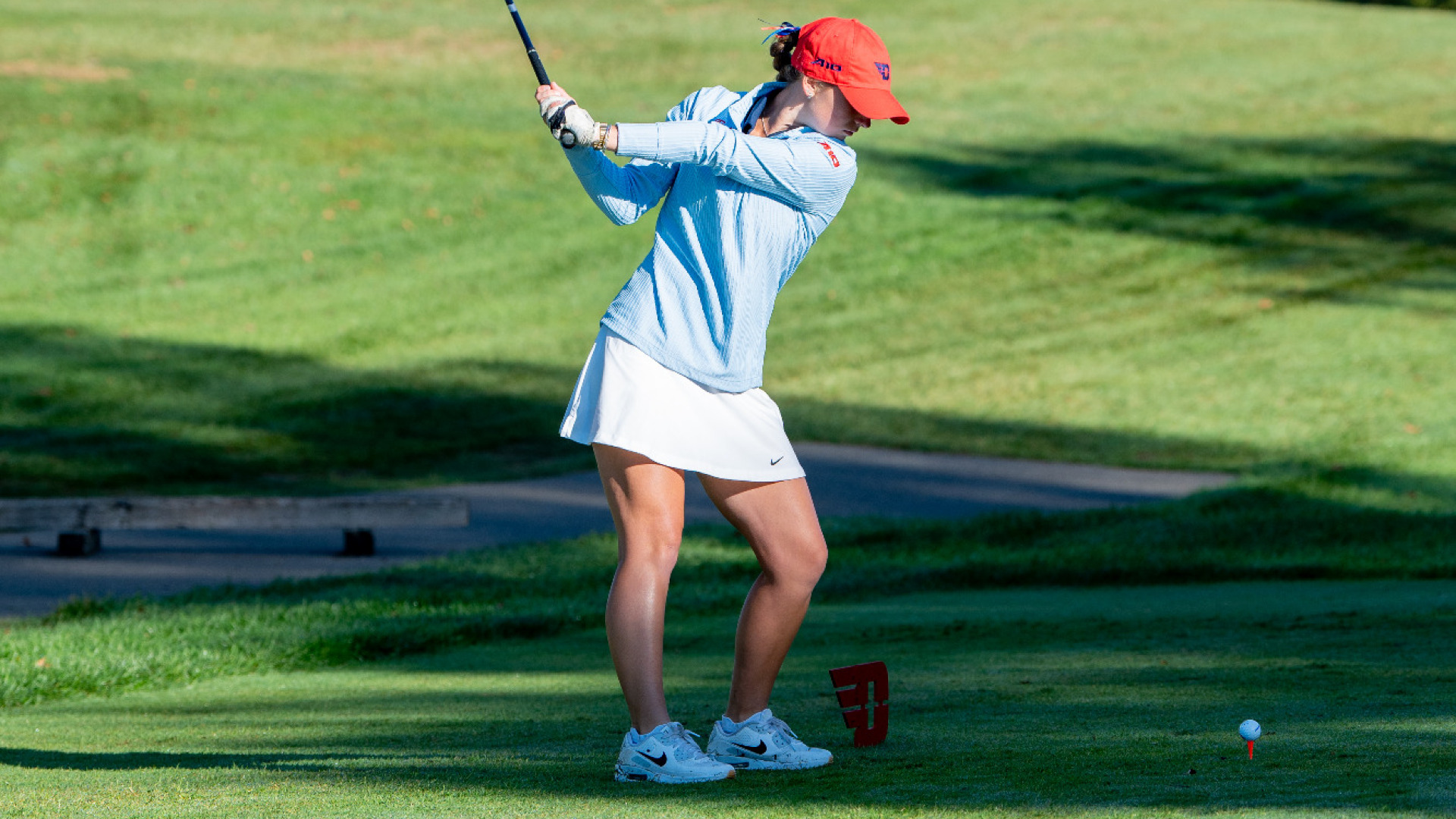 Women's golfer Etta Huffman, wearing a light-blue long-sleeve top, white skort and red hat, with the club above her head at the top of her backswing in a tee box
