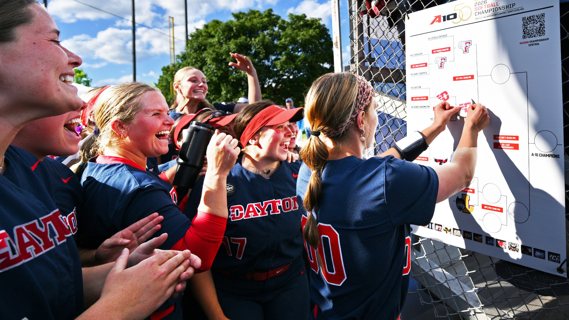 Several members of the softball team wearing navy jerseys, stand behind Camrynn Linneman cheering and smiling as she adds a red winger D sticker to the 2026 Atlantic 10 Championship bracket after defeating Saint Louis