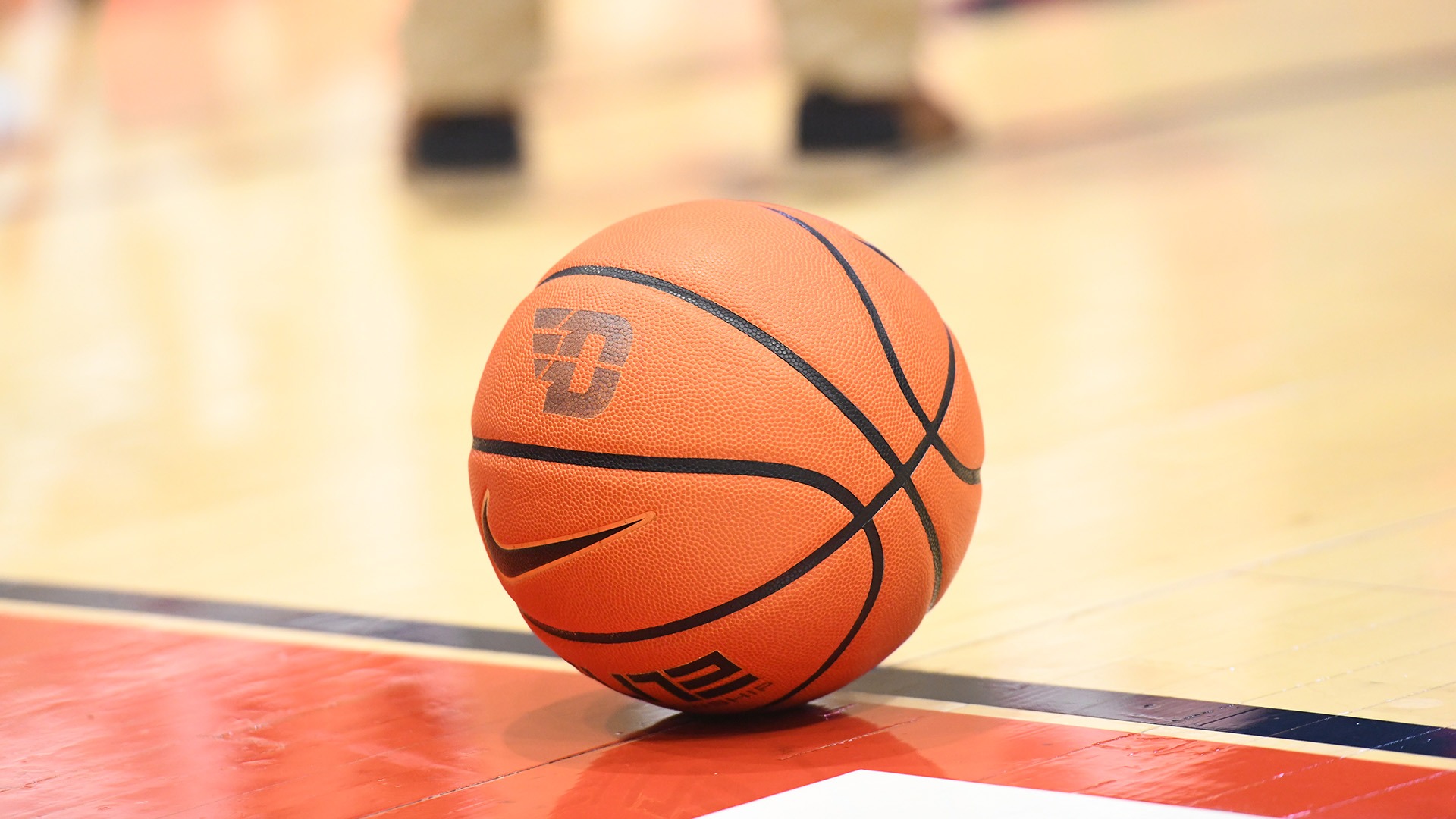 Close-up of an orange Dayton-branded Nike basketball resting on the hardwood near the sideline of a basketball court. The ball is in sharp focus, while the legs and shoes of players in the background are blurred, creating a shallow depth-of-field effect. The court’s red boundary area and polished wood floor are visible beneath the ball.