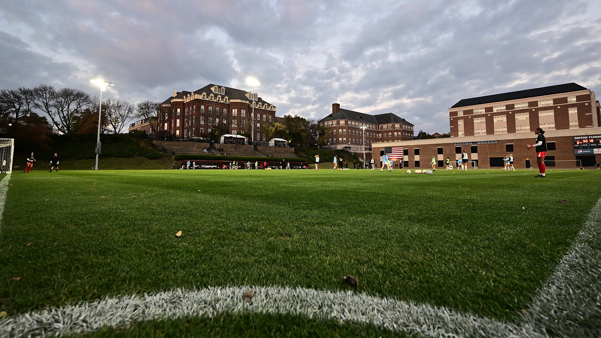 Wide-angle view of the University of Dayton soccer practice field at dusk, with players spread across the pitch during training under the lights. Academic buildings overlook the field in the background beneath a dramatic, cloud-filled evening sky, while the white corner line and green turf dominate the foreground.