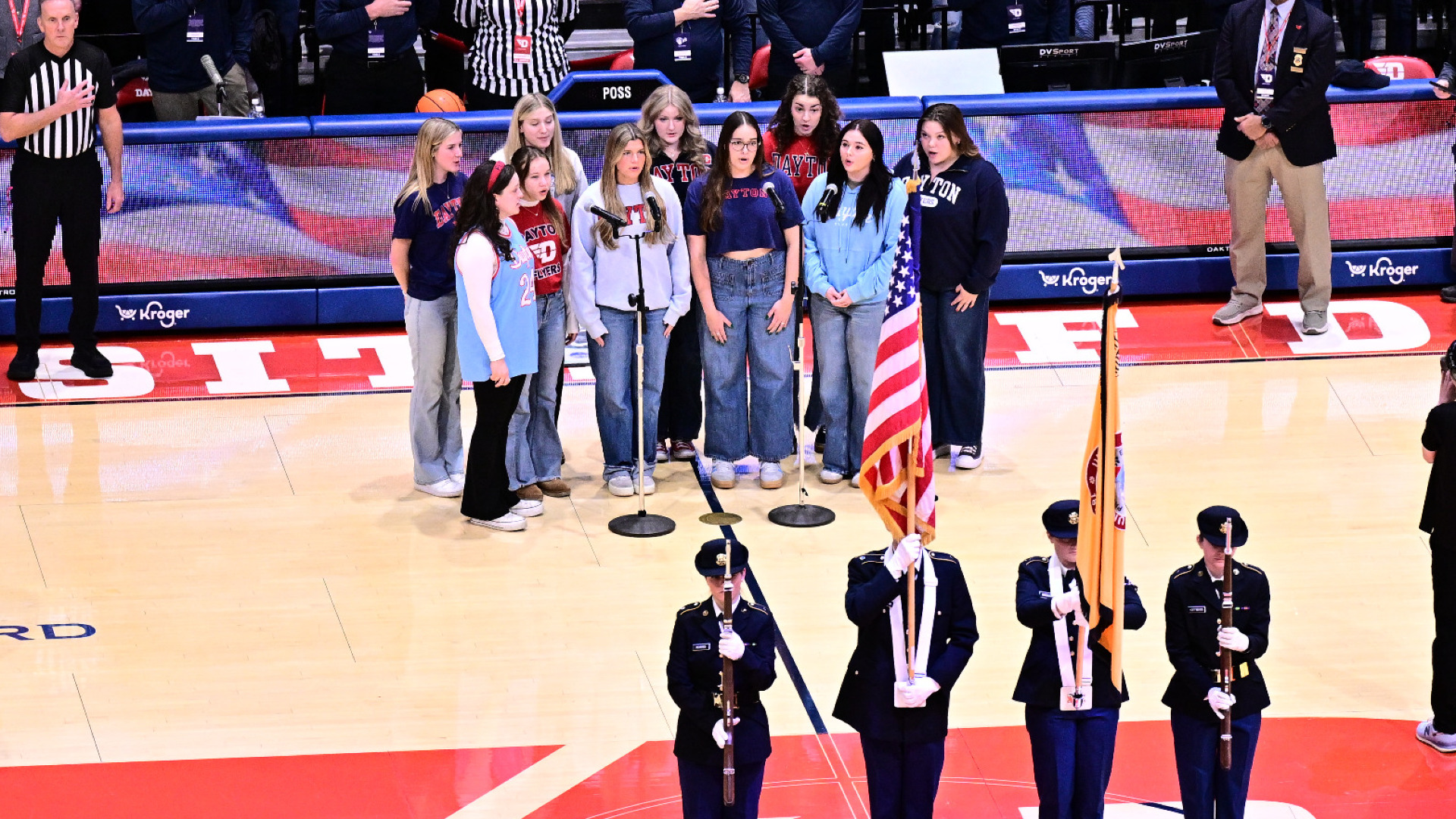 Several members of an acapella singing group, standing behind a colorgaurd on the court, sing the national anthem before a men's basketball game