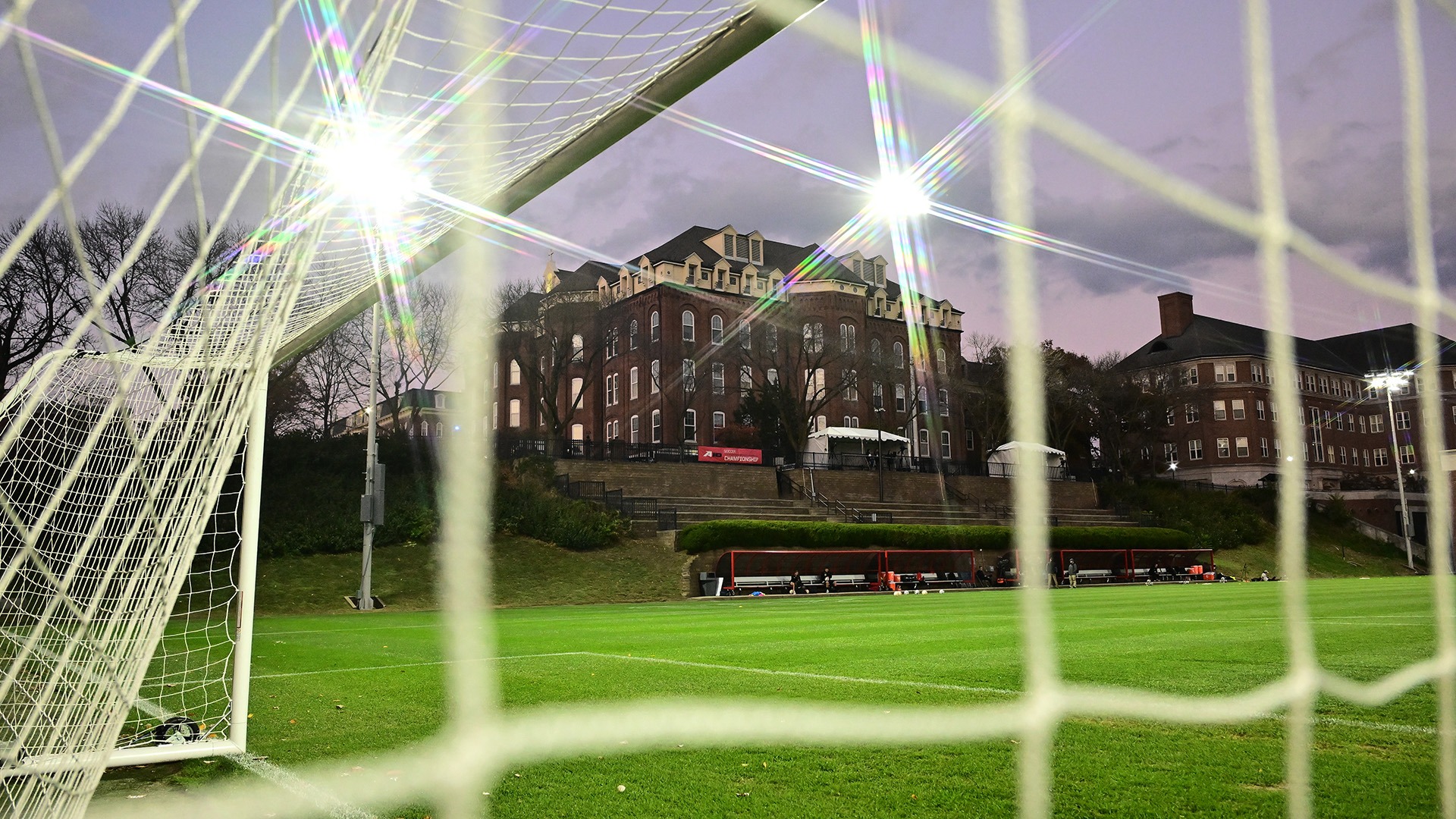 A view from inside a soccer goal looks across a brightly lit soccer field at dusk. The white goal net frames the foreground, while stadium lights shine overhead. Brick campus buildings rise on a hill beyond the field, with team benches along the sideline beneath a cloudy evening sky.
