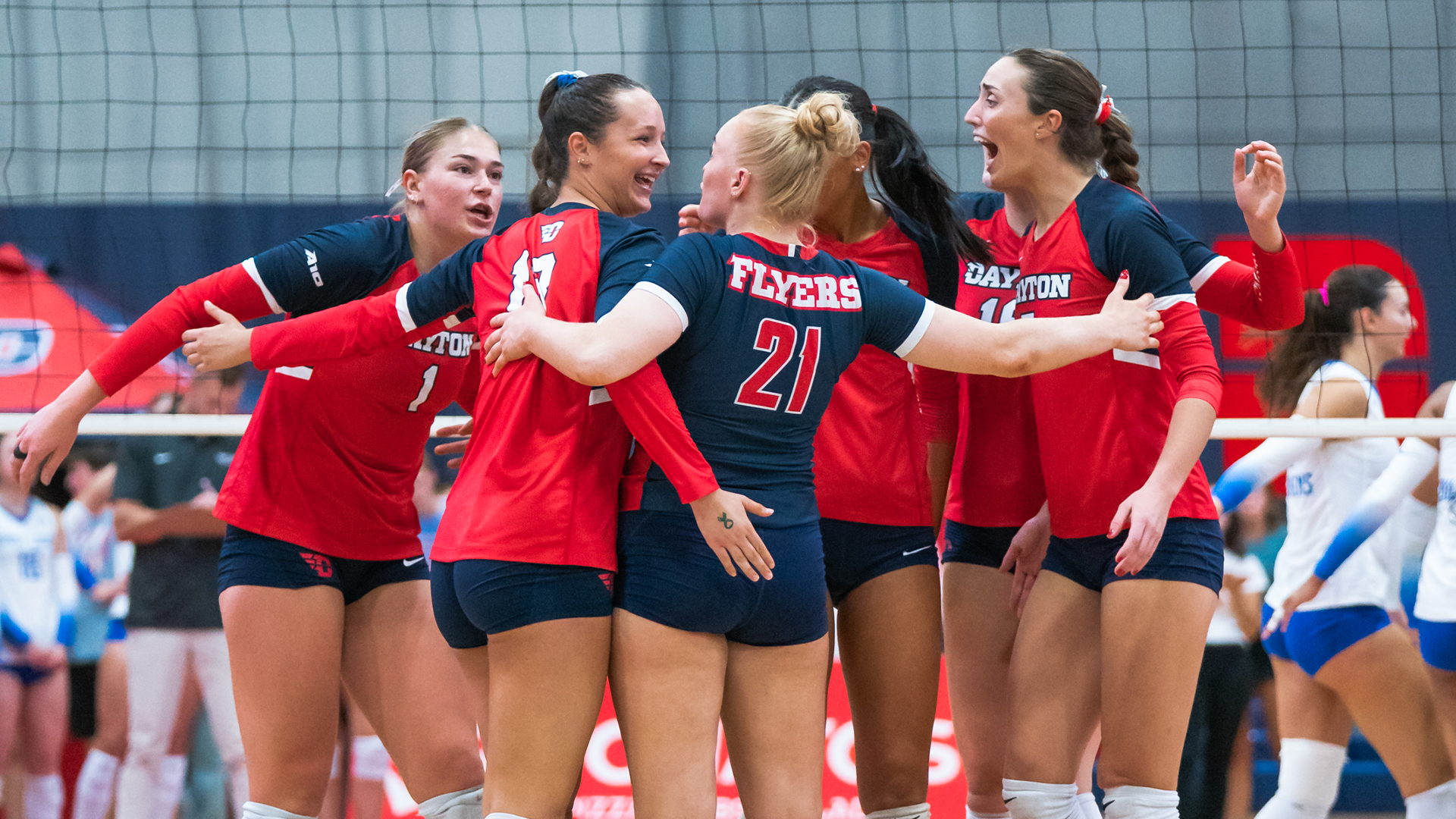 The group of players on the court hug in celebration after a point. Five of them are wearing a red jersey, while the libero wears a navy jersey.
