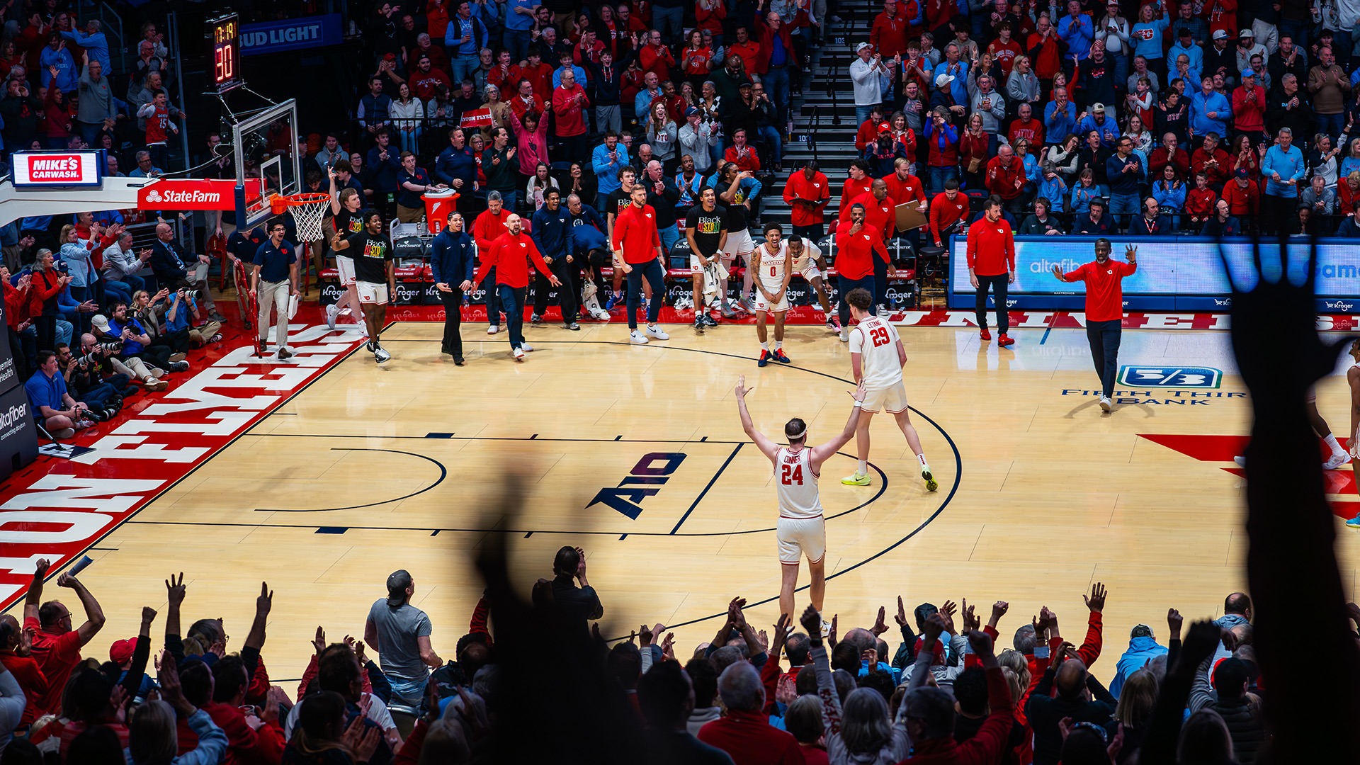 A wide-angle view of a packed basketball arena captures the home crowd celebrating as two players in white uniforms stand on the court with their arms raised toward the fans. Teammates and coaches along the bench leap to their feet in celebration, while spectators throughout the arena cheer and applaud, creating a high-energy atmosphere during the game.
