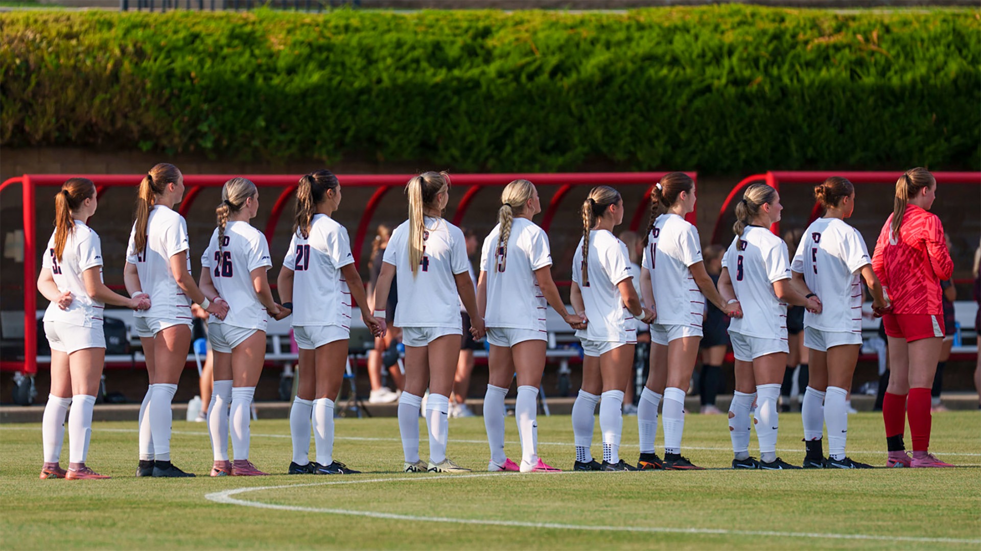Dayton women’s soccer players stand side by side holding hands on the field before a match, wearing white uniforms with the goalkeeper in red.