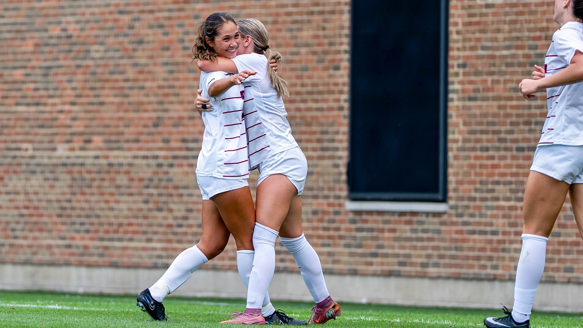 Dayton women’s soccer player Olivia Baca embraces a teammate and smiles in celebration on the field as a teammate approaches.