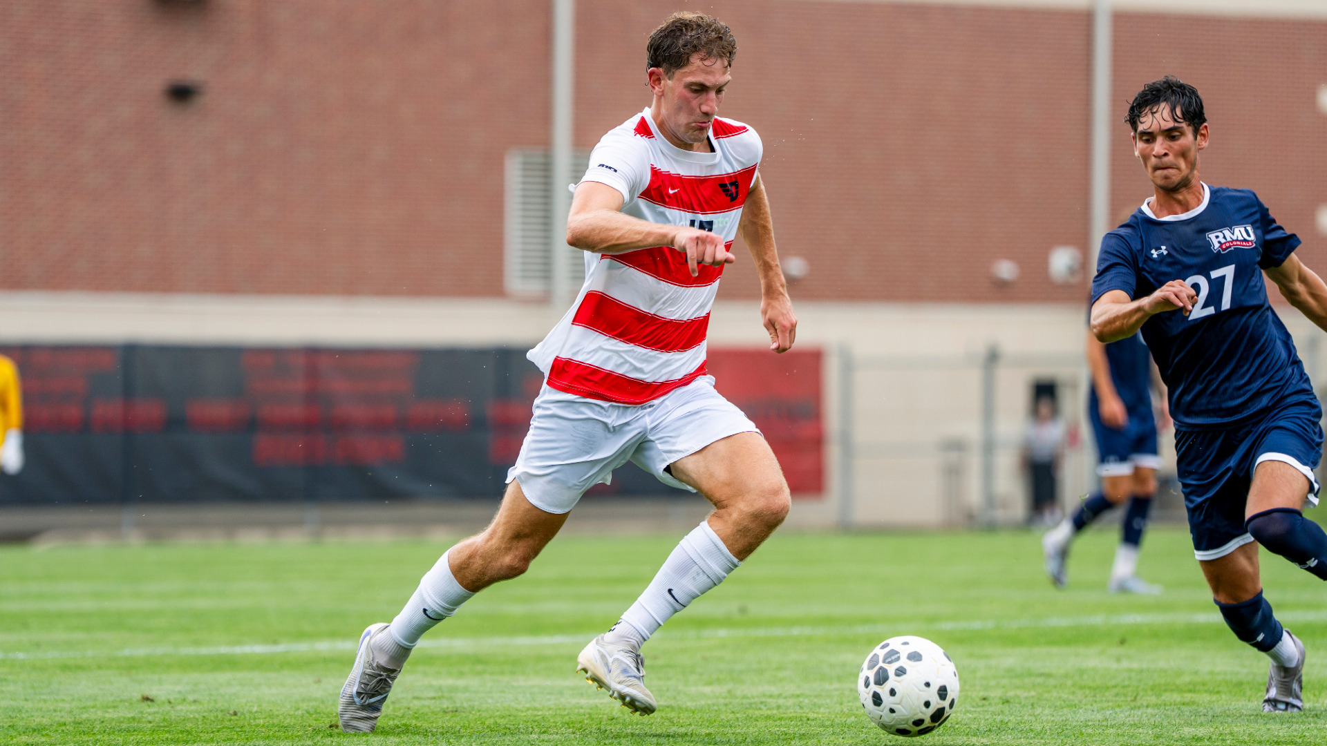 Men's soccer player Mile Bonham, wearing a red and white striped jersey, white shorts and white socks, dribbles a ball past a Robert Morris defender during a game at Baujan Field