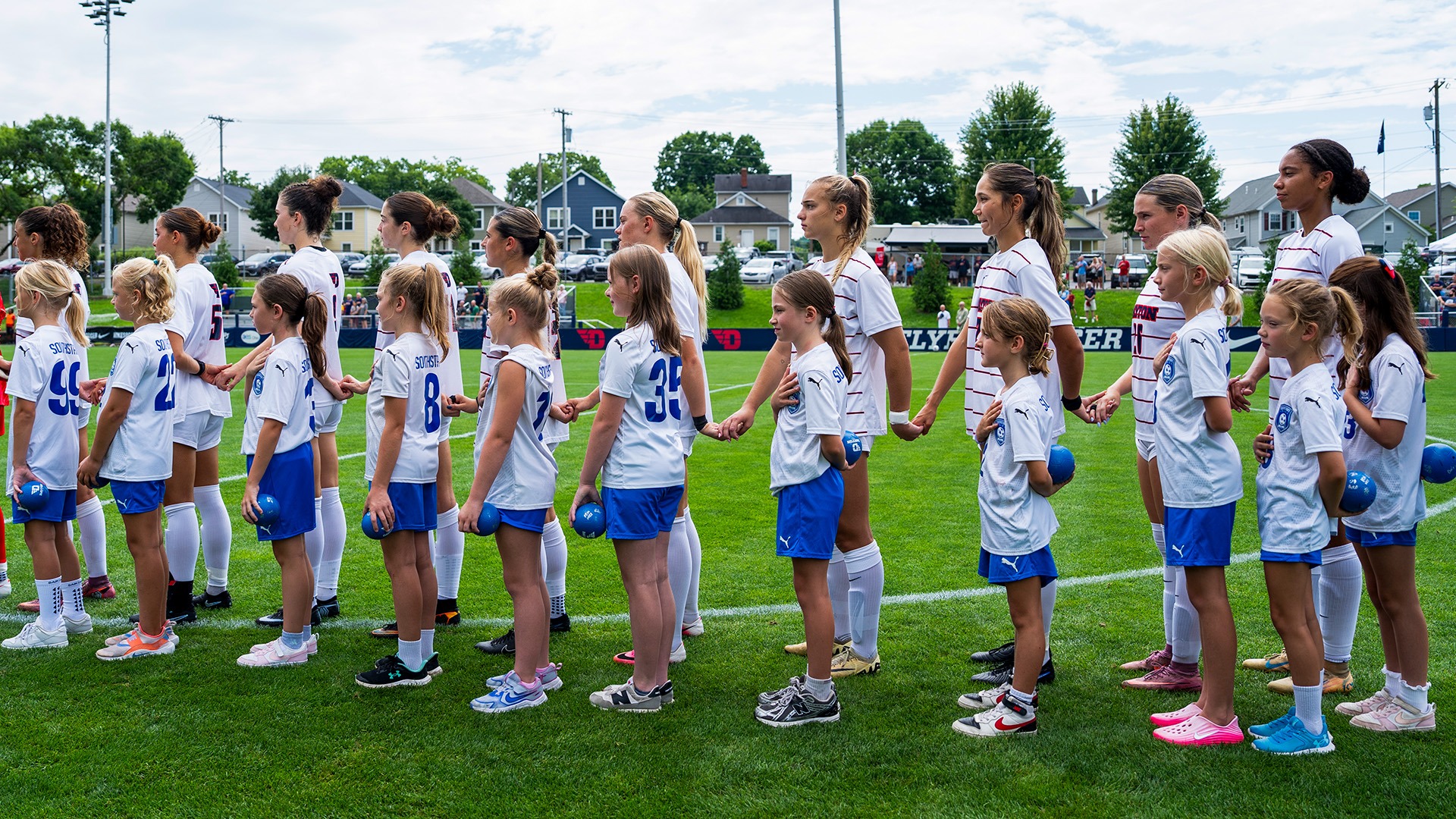 Women’s soccer players stand hand-in-hand with young girls on the field during pregame introductions.