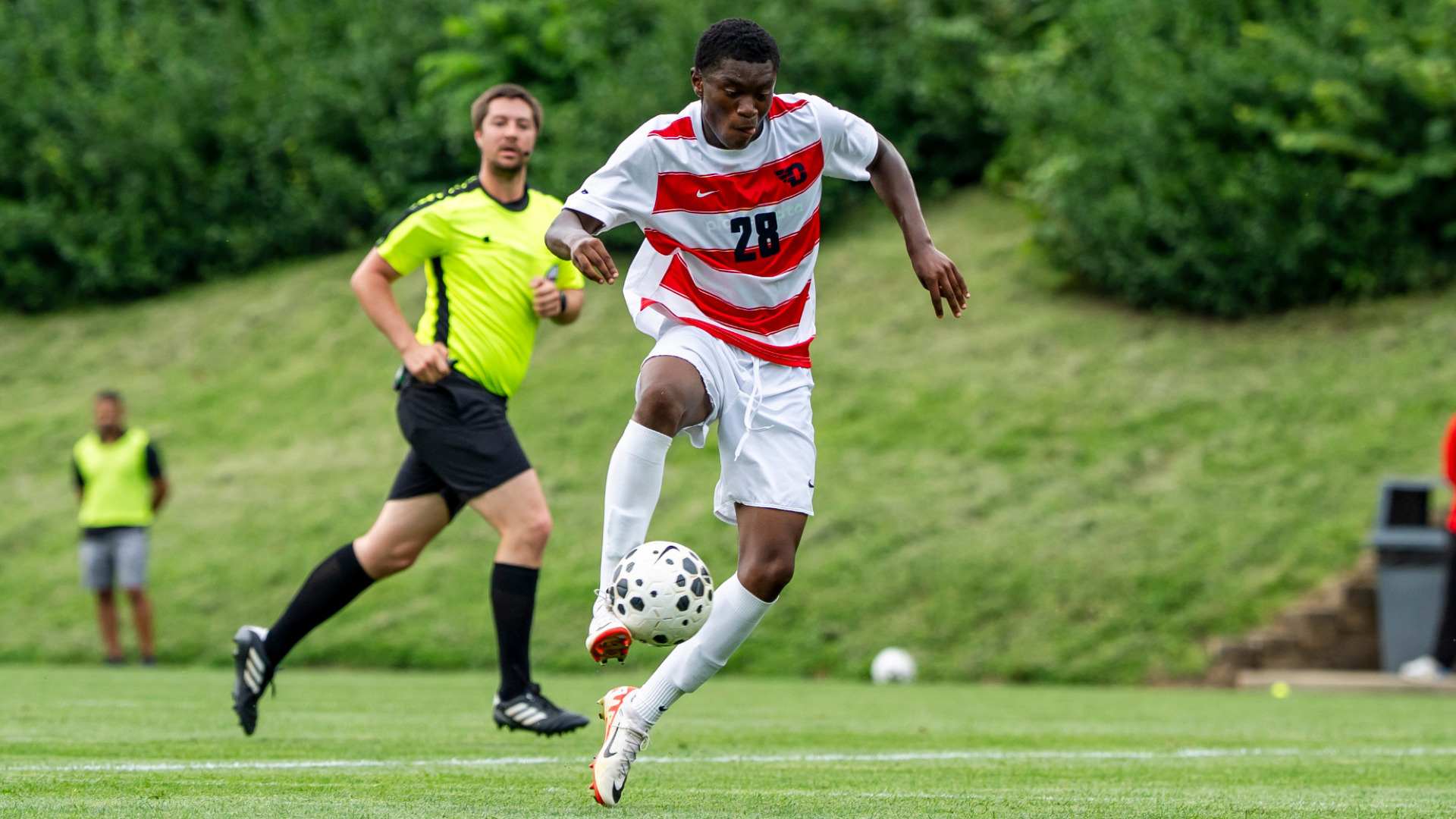 Men's soccer player Uri Koffi, wearing a red and white jersey with white shorts and white socks, receives the ball midfield during a match at Baujan Field