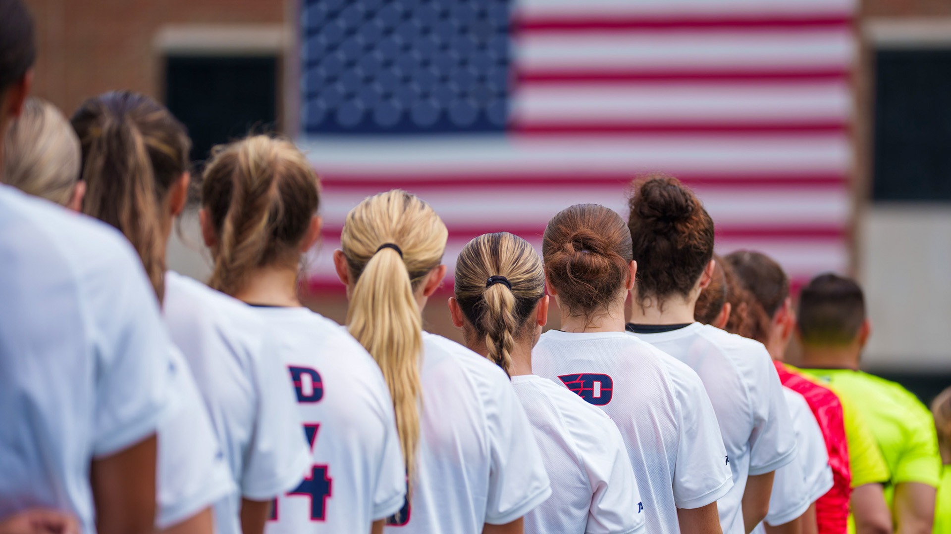 Dayton women’s soccer players stand in a line facing a large American flag before a match.