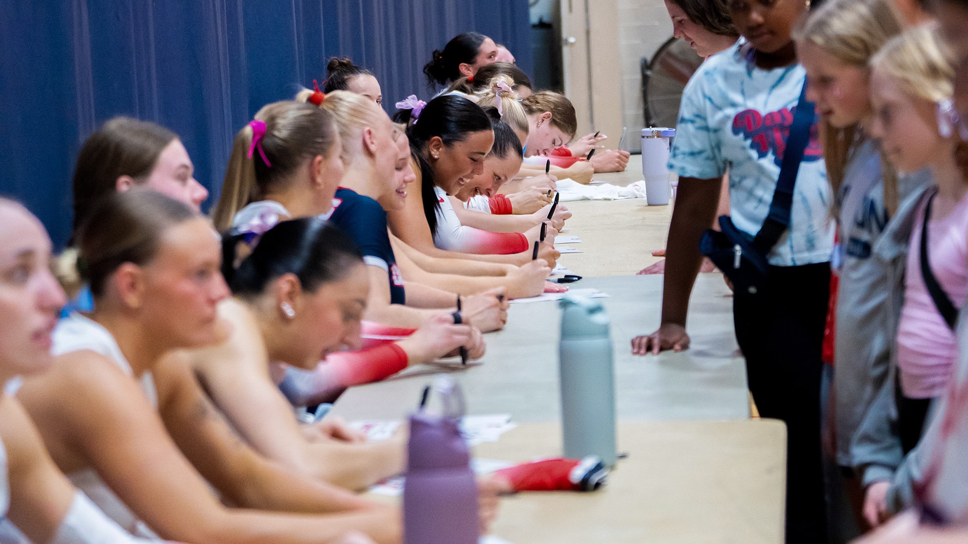 The 2025 volleyball team signs autographs for young fans. The players are sitting on the left side of the pictures, wearing white uniforms. The young girls are standing on the right side of the table.