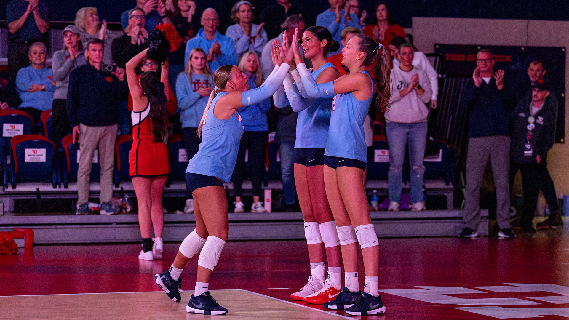 Lily Barry high-fives Abigail Cresse and Kamryn Hunt during starting lineups. The lights in the Frericks Center are down, and the players are wearing Chapel Blue uniforms.